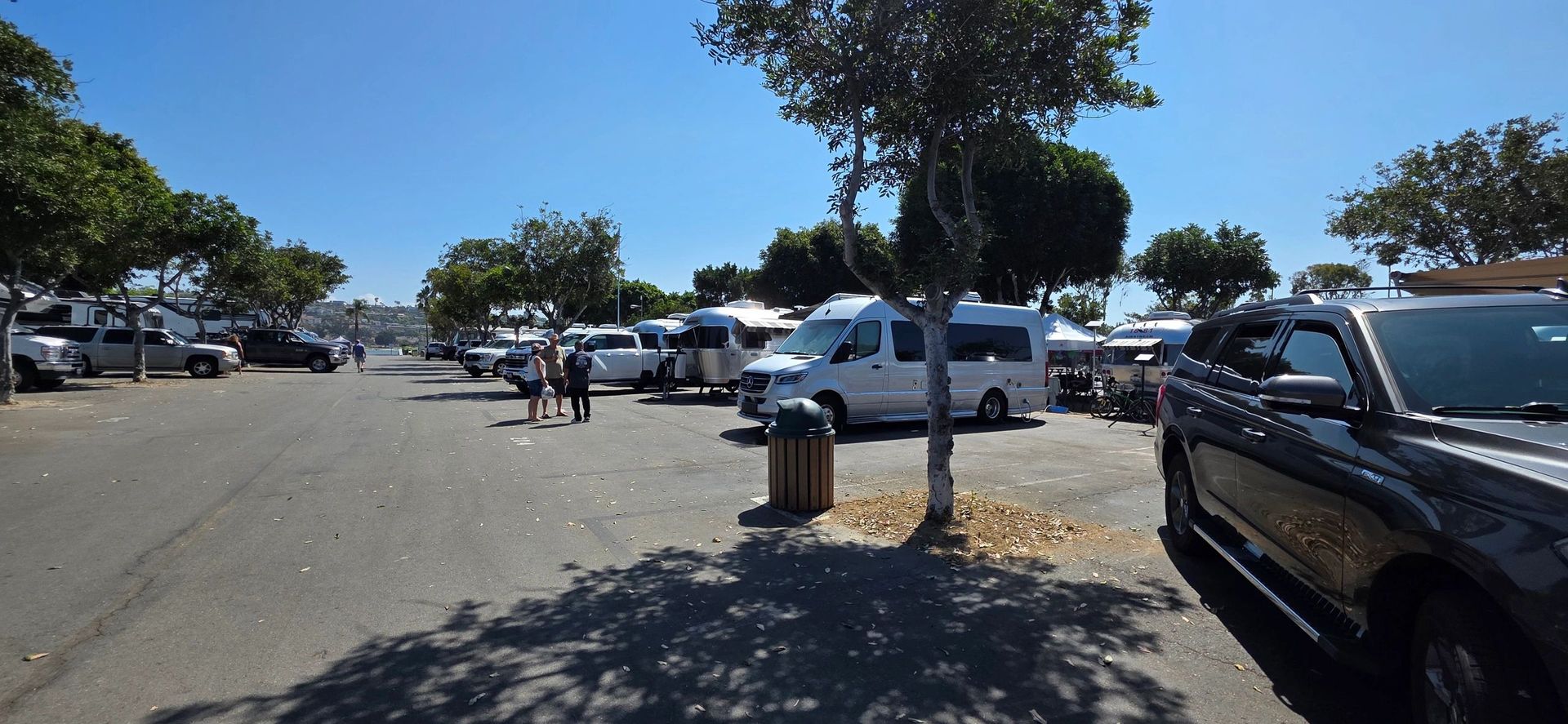 A parking lot with vehicles, trees, and people under a bright blue sky.