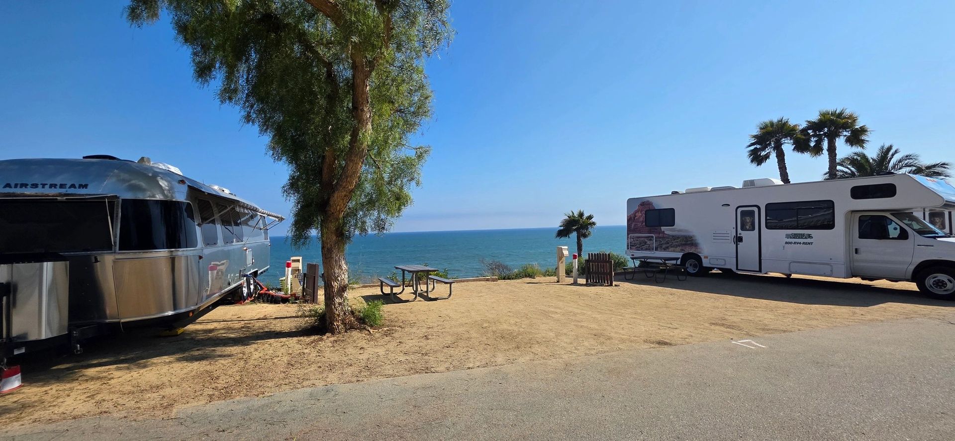 An Airstream trailer and a motorhome parked at a beach campsite, with a view of the ocean.