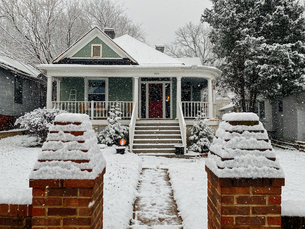 A house with a bricked entryway with trees in its yard all covered in snow during a snowstorm.