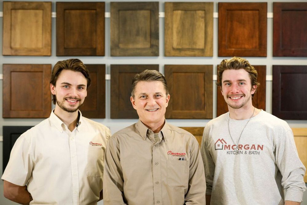 Three men smiling in front of a wall of wooden cabinet door samples. The men wear work shirts with a company logo.