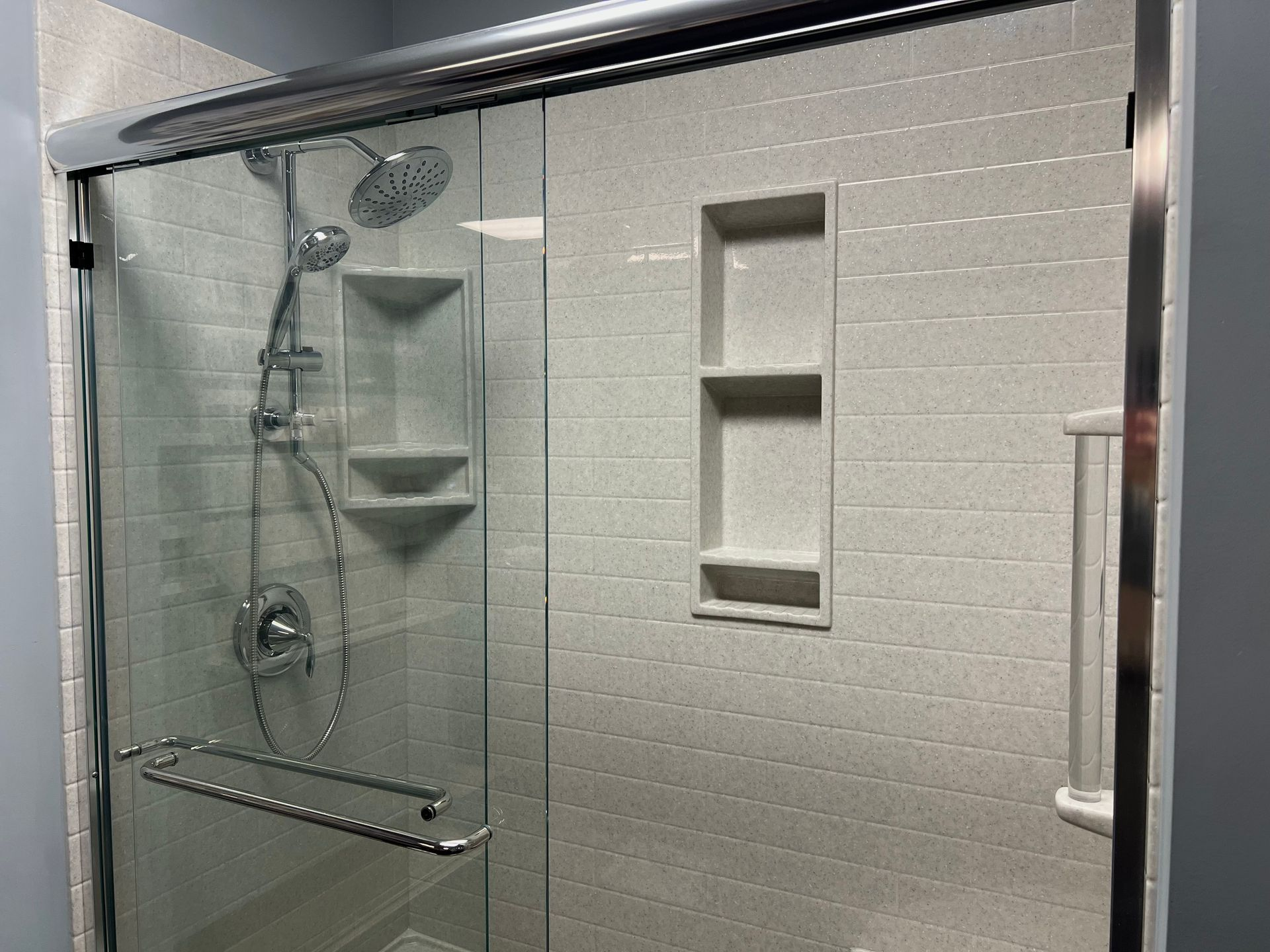 Shower with glass sliding door and built-in shelves. The walls are light-colored with a textured pattern. Chrome fixtures.