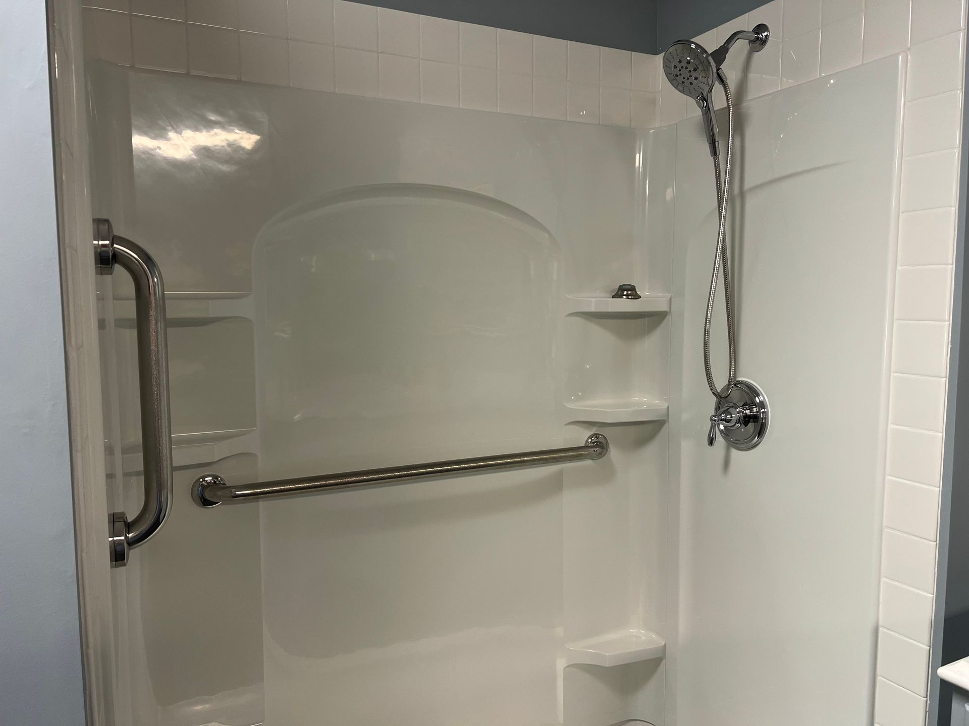 Accessible white shower with grab bars, shelves, and handheld showerhead. Blue walls surround the shower.
