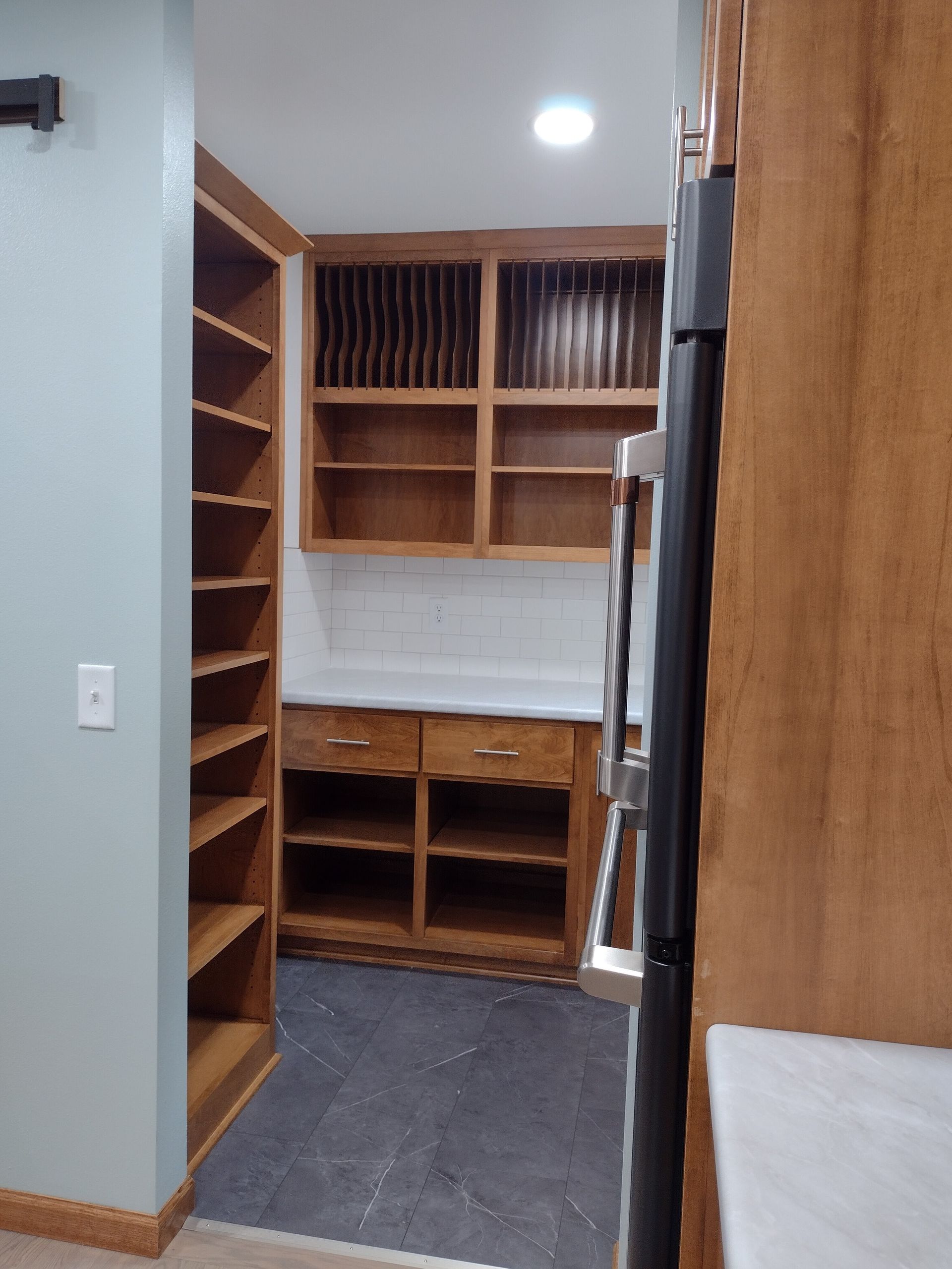 A pantry with wooden shelves and cabinets. The walls are painted light blue. A stainless steel refrigerator is visible on the right.