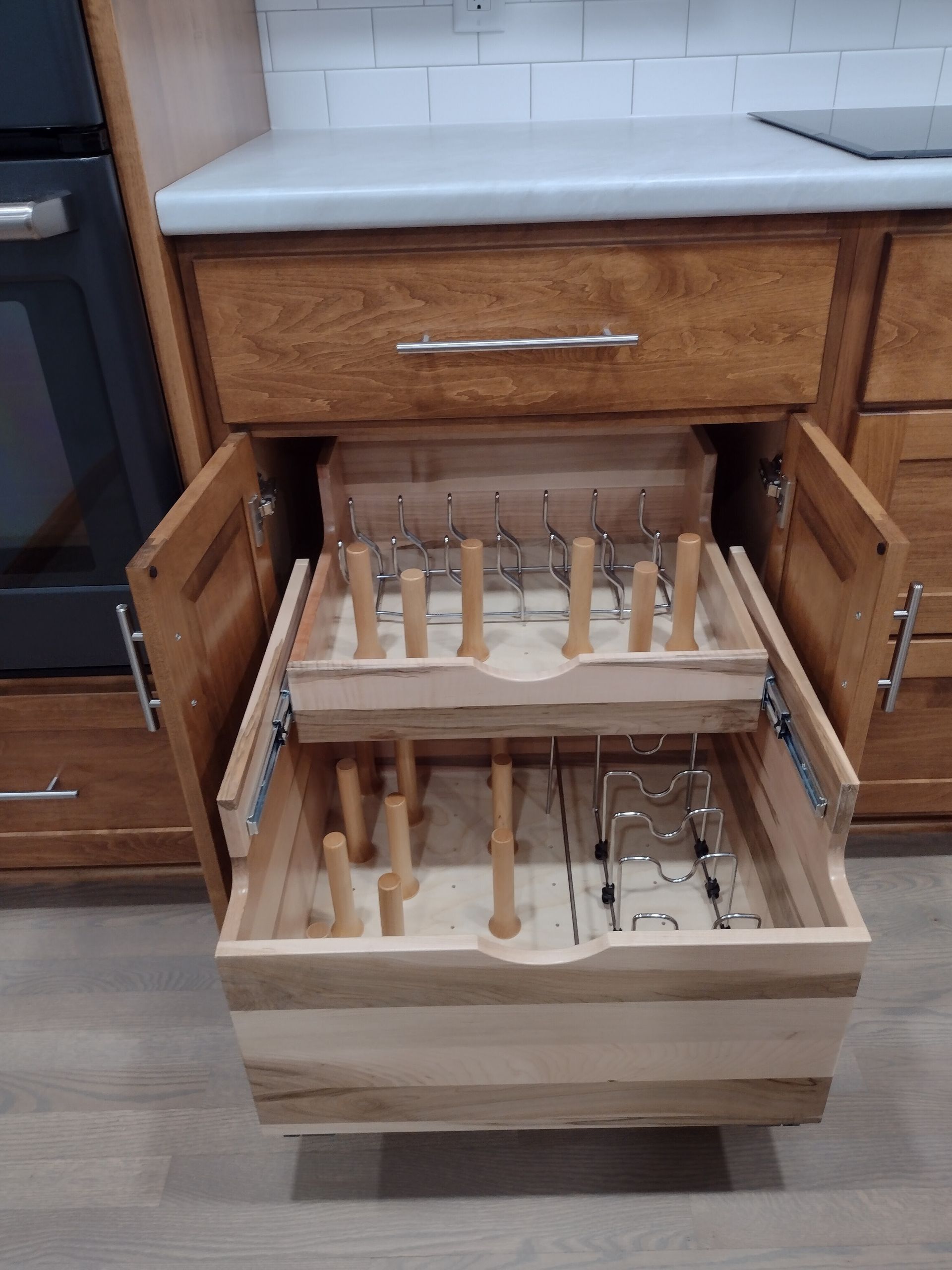 Wooden kitchen cabinet with an open drawer revealing a custom-built stemware storage system with multiple pegs and metal racks.