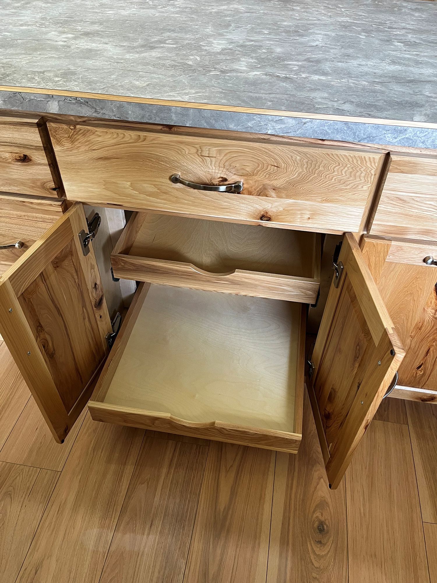Wooden kitchen cabinet with open doors and pull-out shelves under a gray countertop.