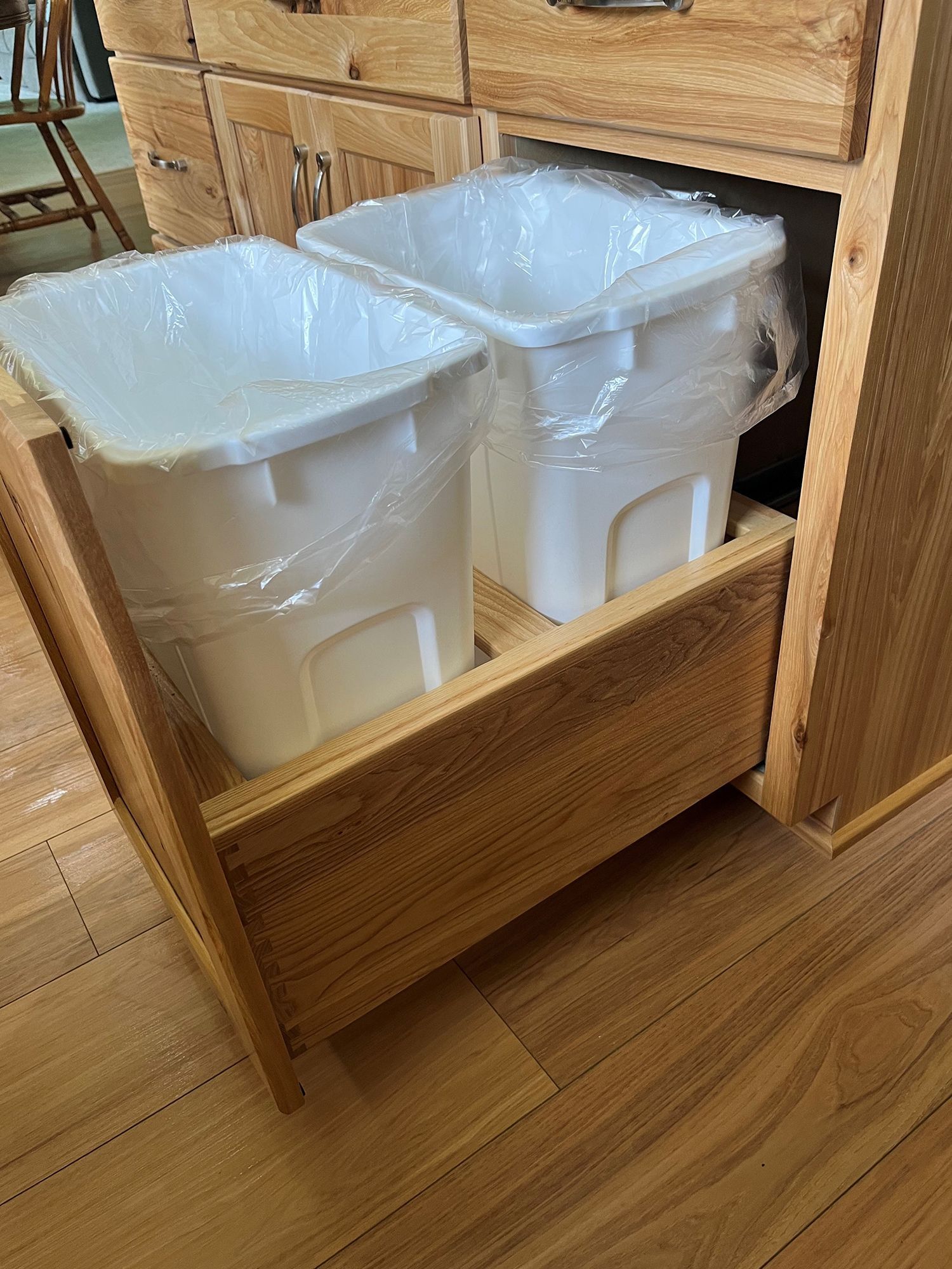 Wooden cabinet with a drawer holding two white plastic waste bins, partially covered with plastic.