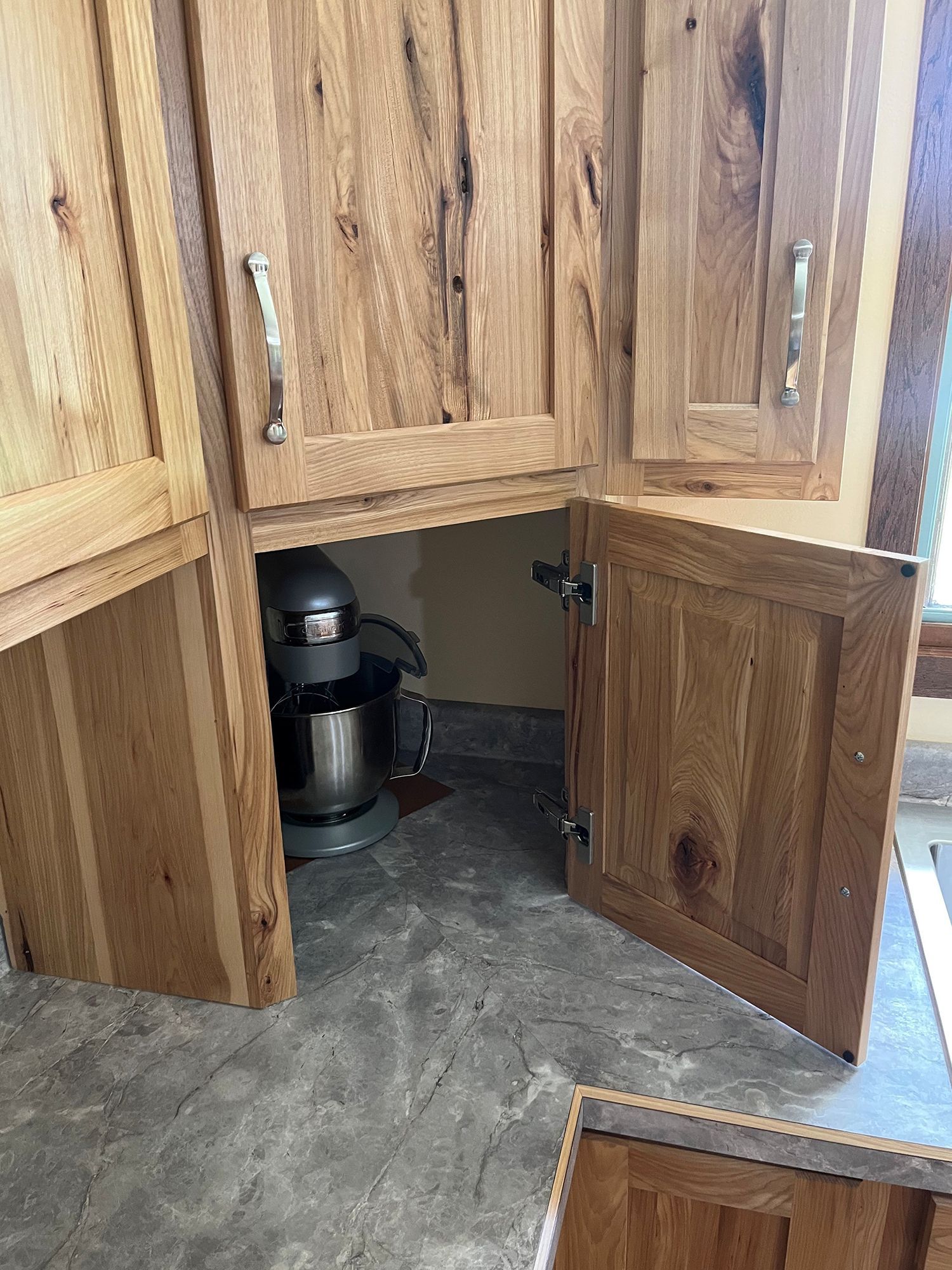 Open wooden kitchen cabinets with a stand mixer inside, on a gray floor.