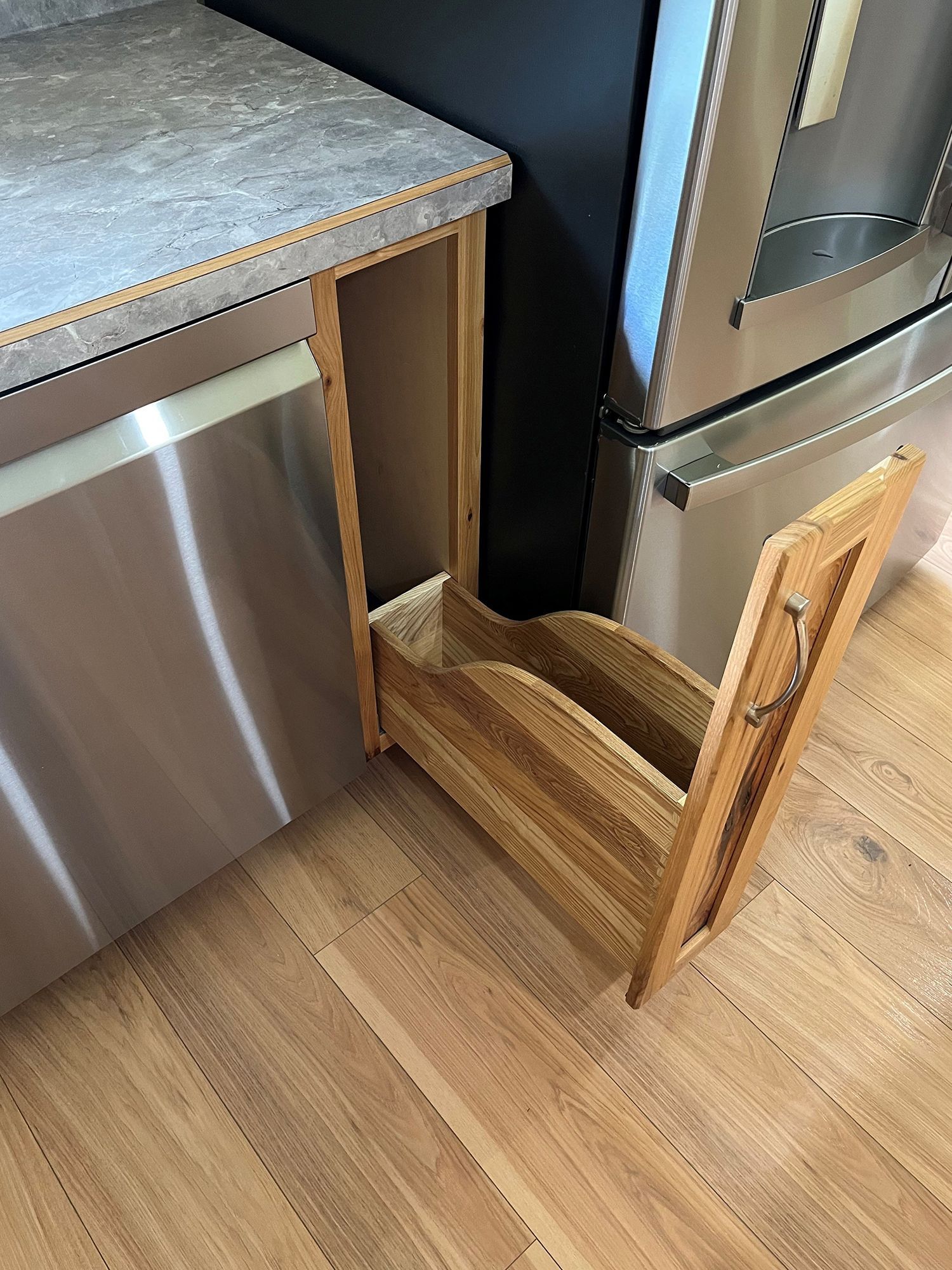 Wooden pull-out cabinet with two curved dividers, next to a stainless steel dishwasher and refrigerator in a kitchen.