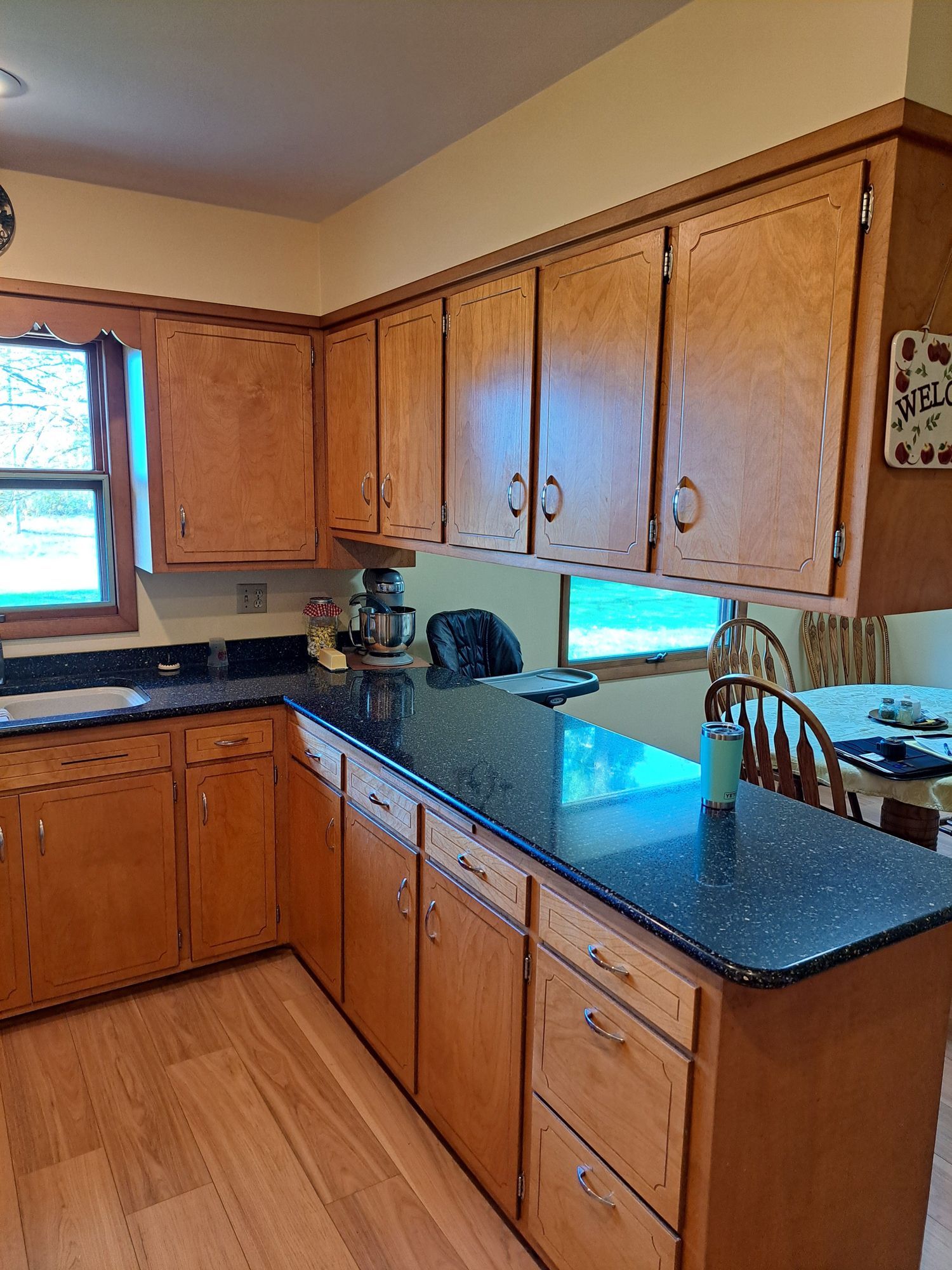 Wooden kitchen cabinets with black countertops. A window provides natural light, and a dining table is partially visible.