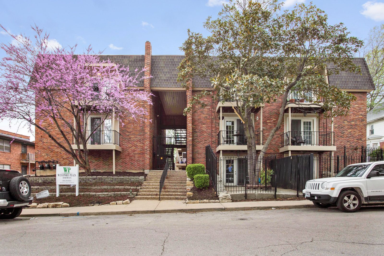 A two-story apartment building with beige siding and dark balconies, surrounded by dormant trees and landscaping.