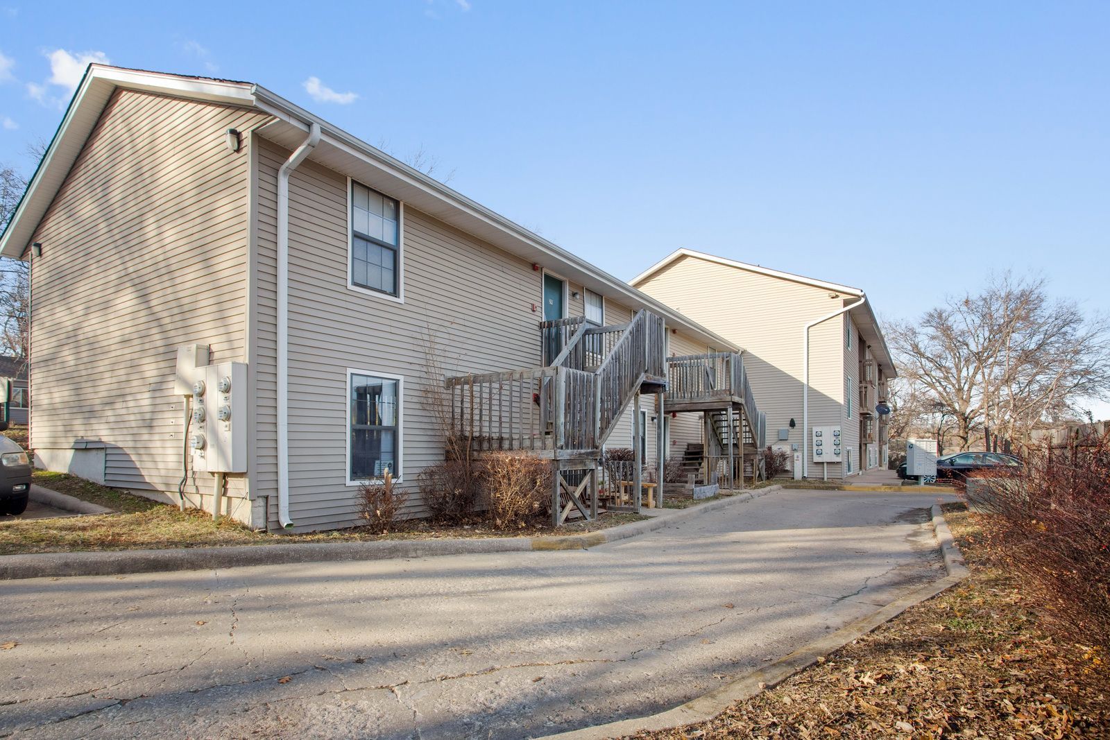A two-story apartment building with beige siding and dark balconies, surrounded by dormant trees and landscaping.