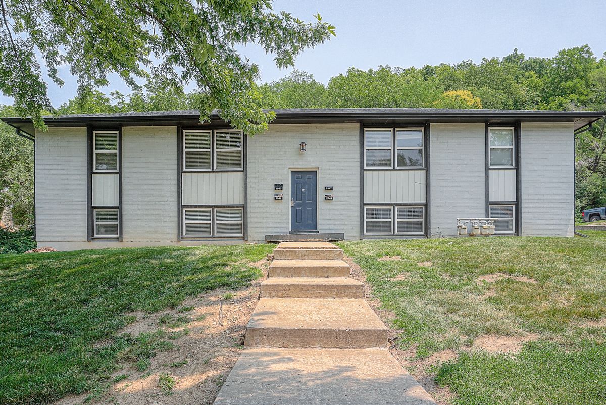 A two-story apartment building with beige siding and dark balconies, surrounded by dormant trees and landscaping.