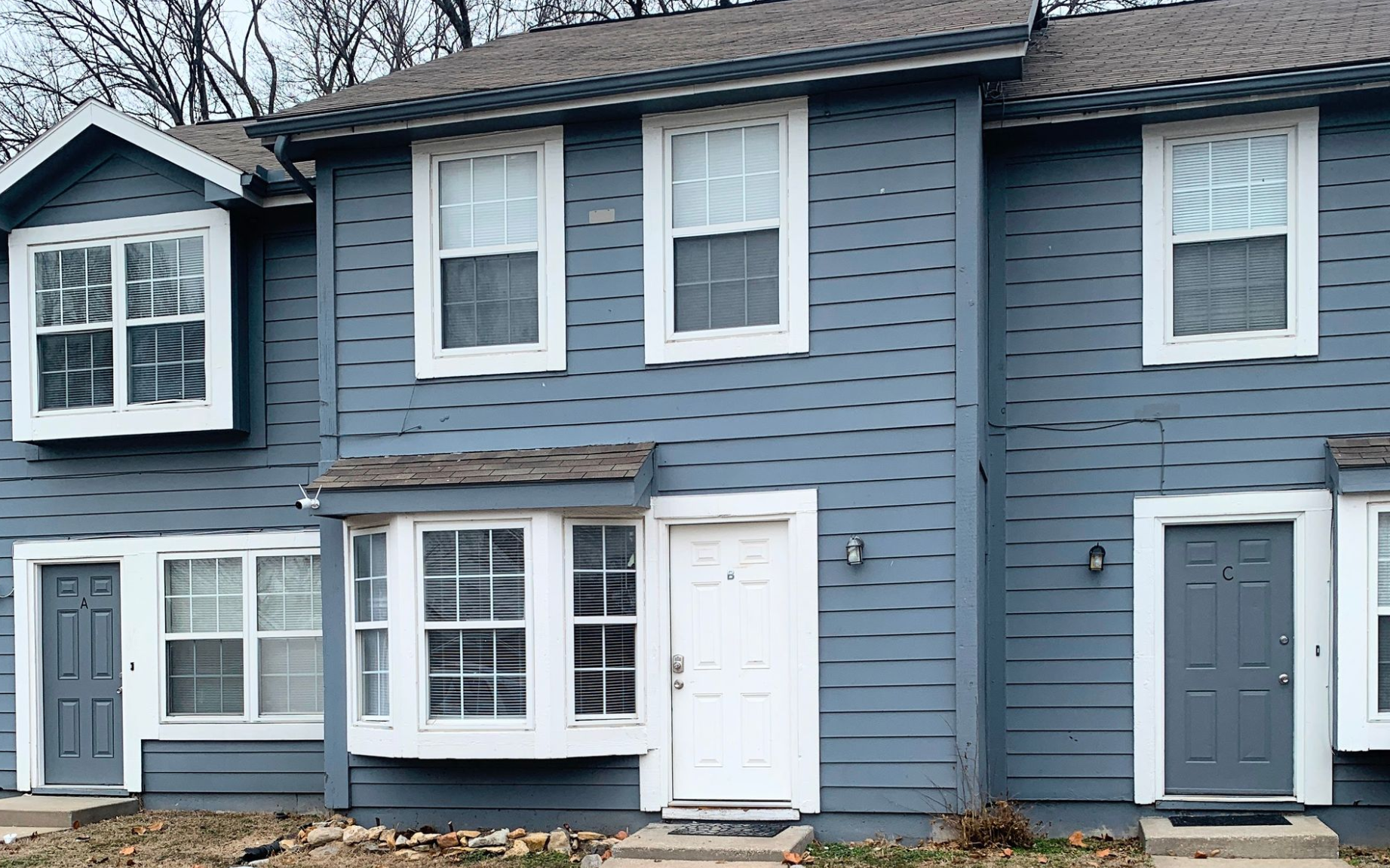 A two-story apartment building with beige siding and dark balconies, surrounded by dormant trees and landscaping.