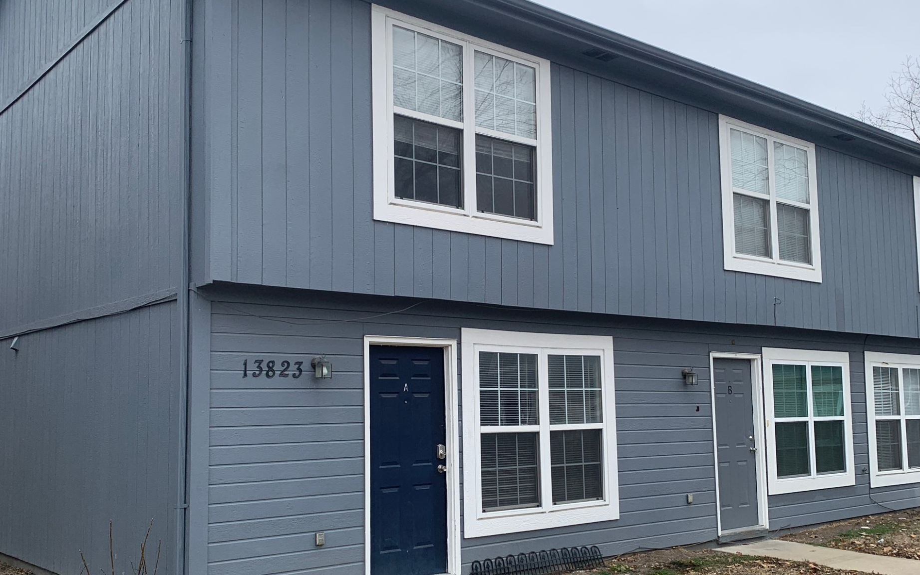 A three-story, red brick apartment building with a recessed center entrance, stone foundation, and a dark overhanging roof.