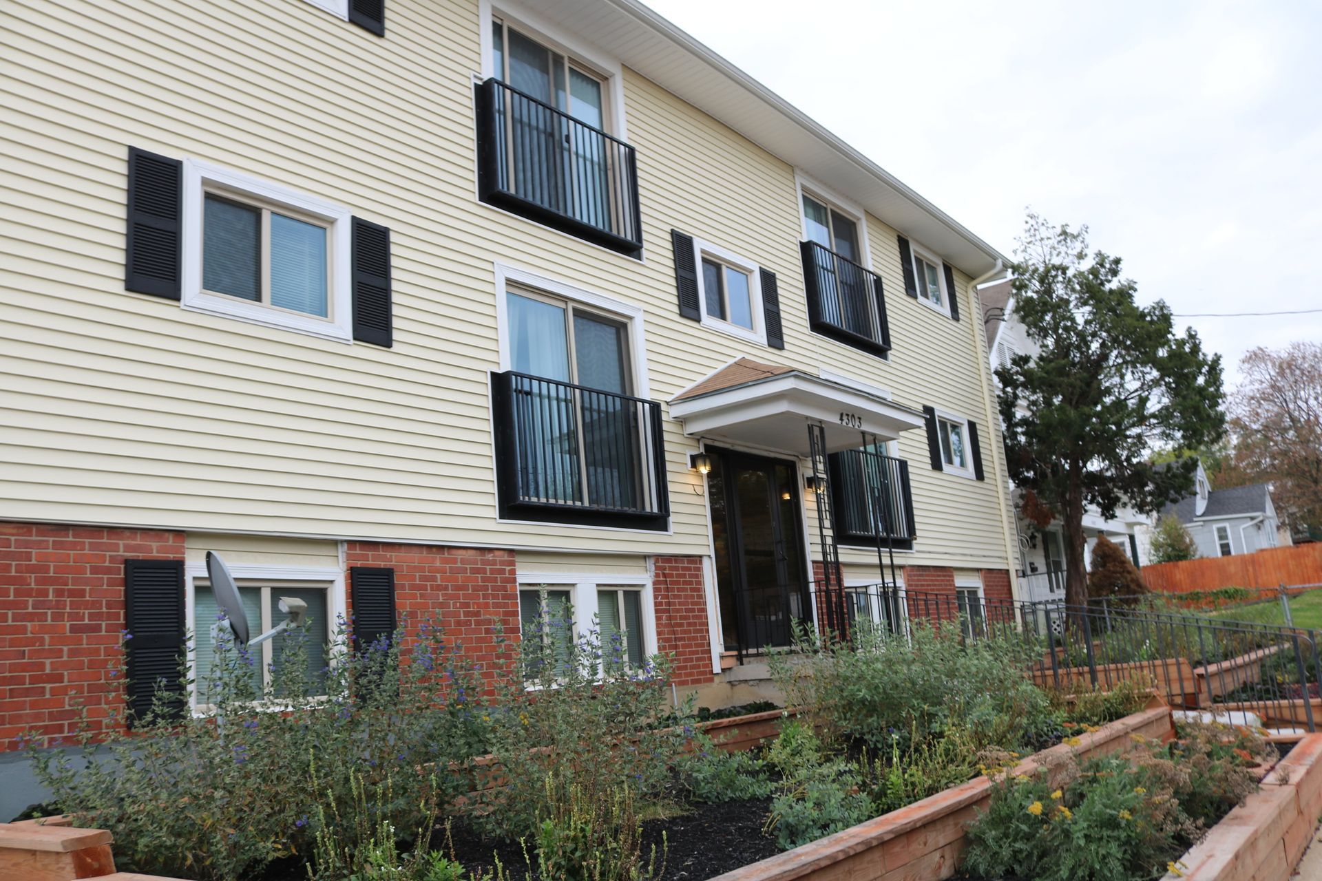 A two-story apartment building with beige siding and dark balconies, surrounded by dormant trees and landscaping.