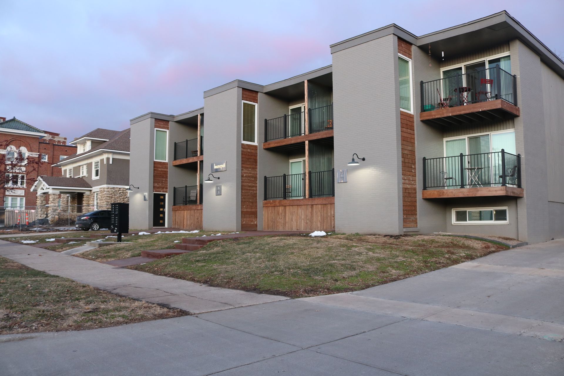 A two-story apartment building with beige siding and dark balconies, surrounded by dormant trees and landscaping.