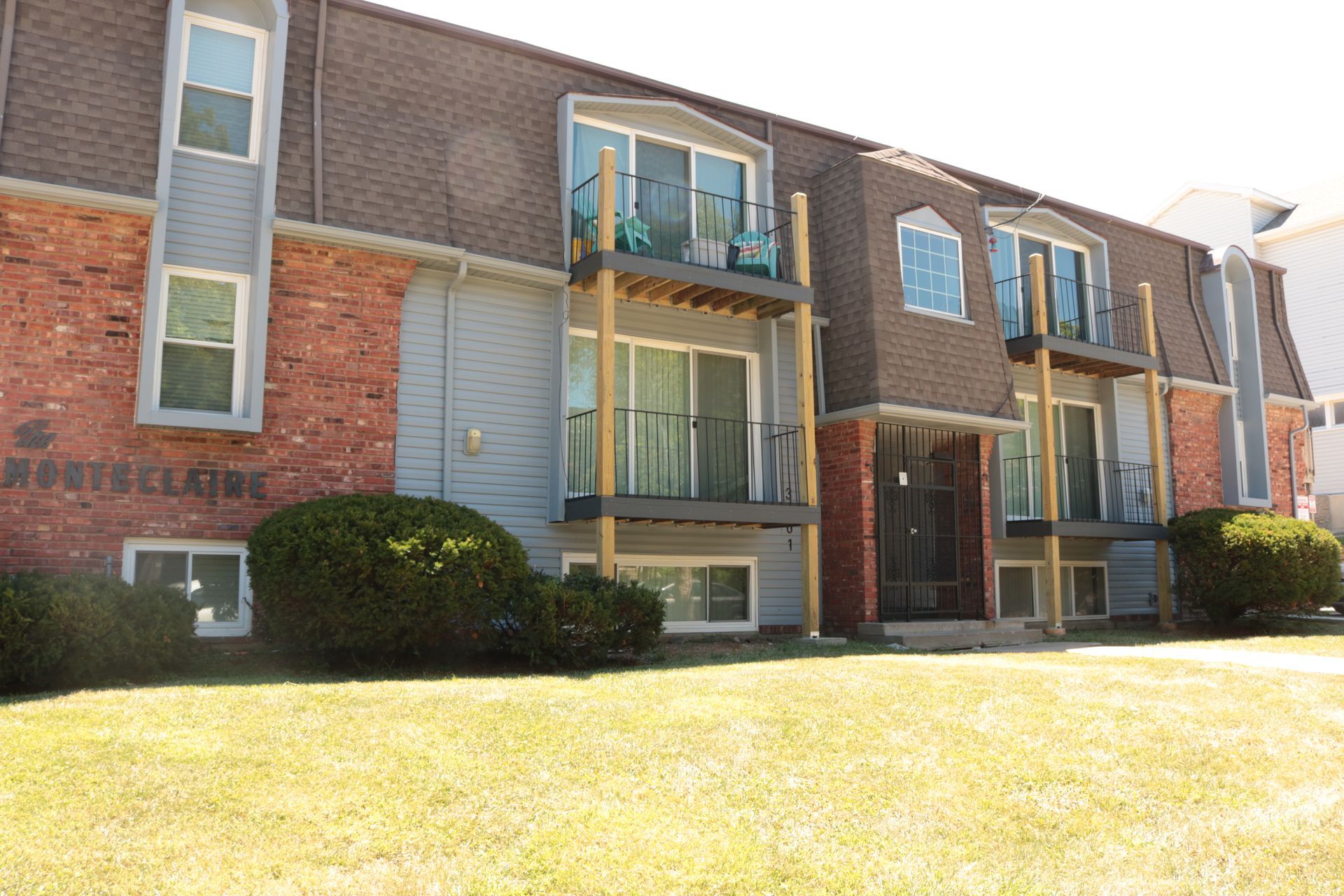 A two-story apartment building with beige siding and dark balconies, surrounded by dormant trees and landscaping.