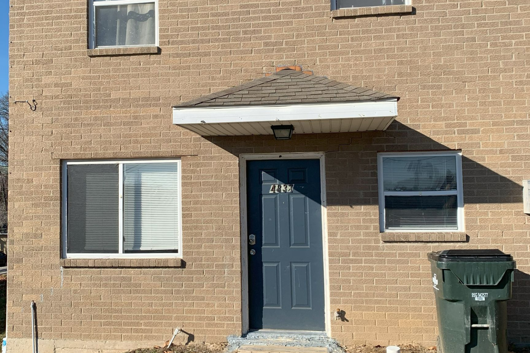 A two-story apartment building with beige siding and dark balconies, surrounded by dormant trees and landscaping.