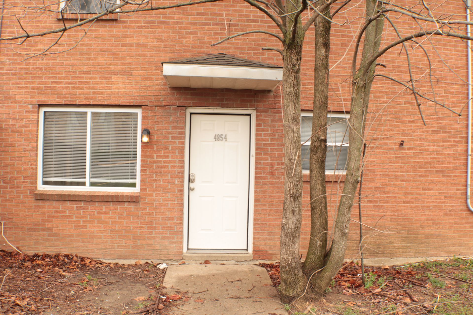 A two-story apartment building with beige siding and dark balconies, surrounded by dormant trees and landscaping.