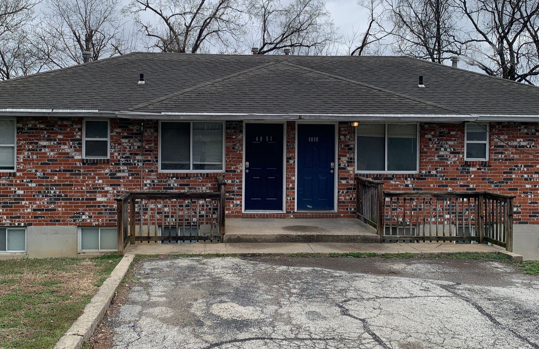 A two-story apartment building with beige siding and dark balconies, surrounded by dormant trees and landscaping.