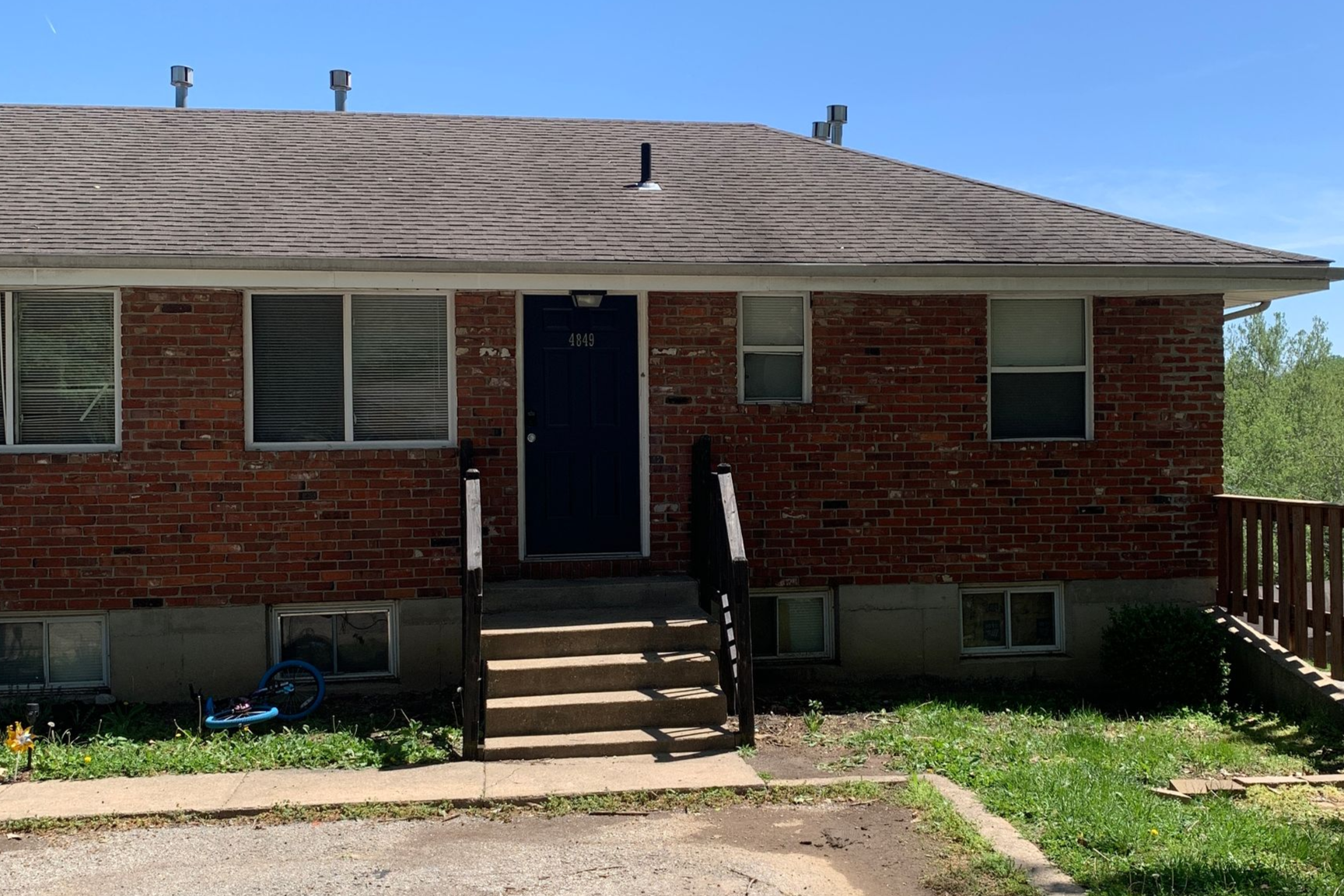 A three-story, red brick apartment building with a recessed center entrance, stone foundation, and a dark overhanging roof.