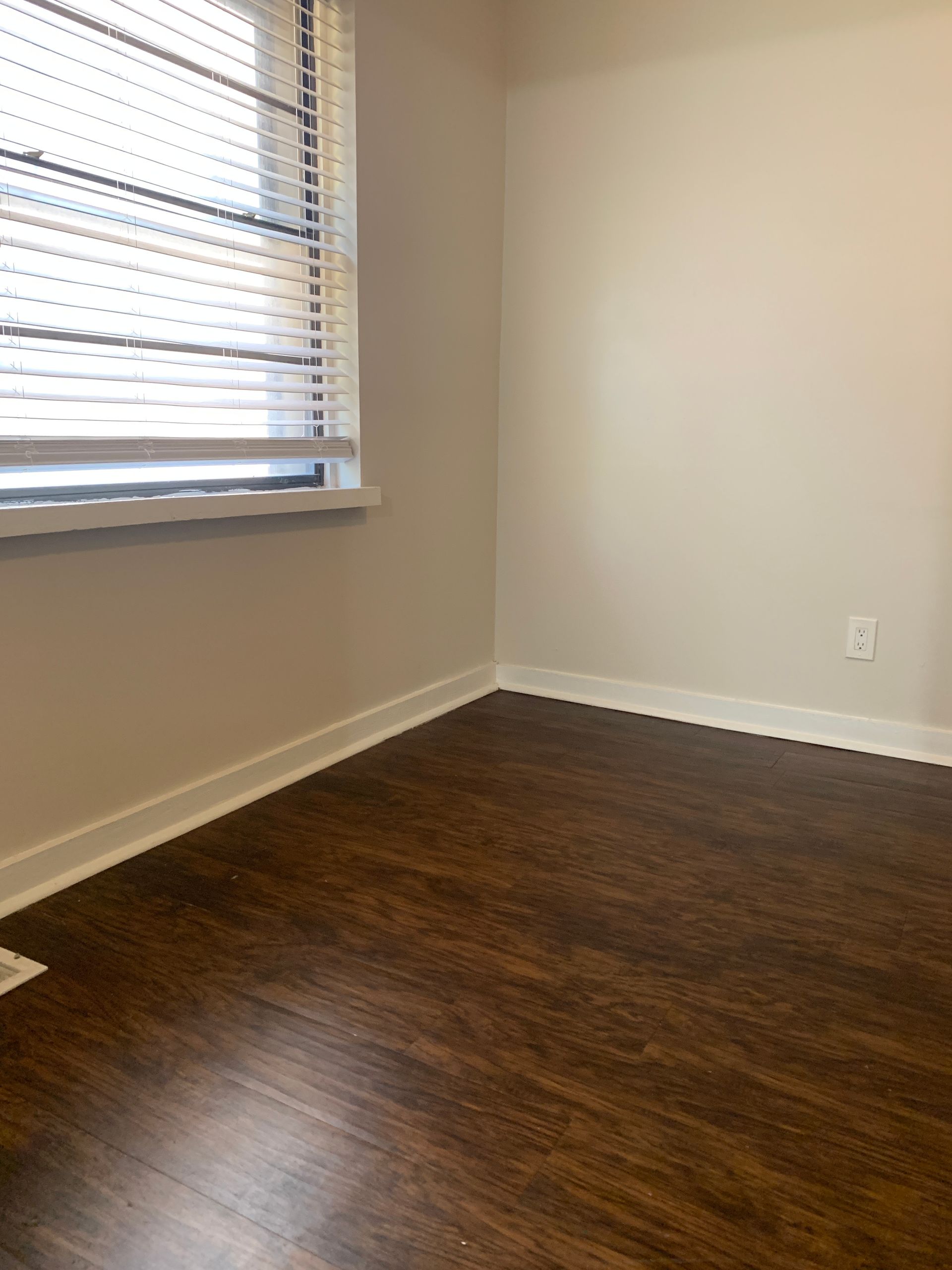 Empty room with dark wood-look flooring, neutral walls, a window with blinds, and a white baseboard.