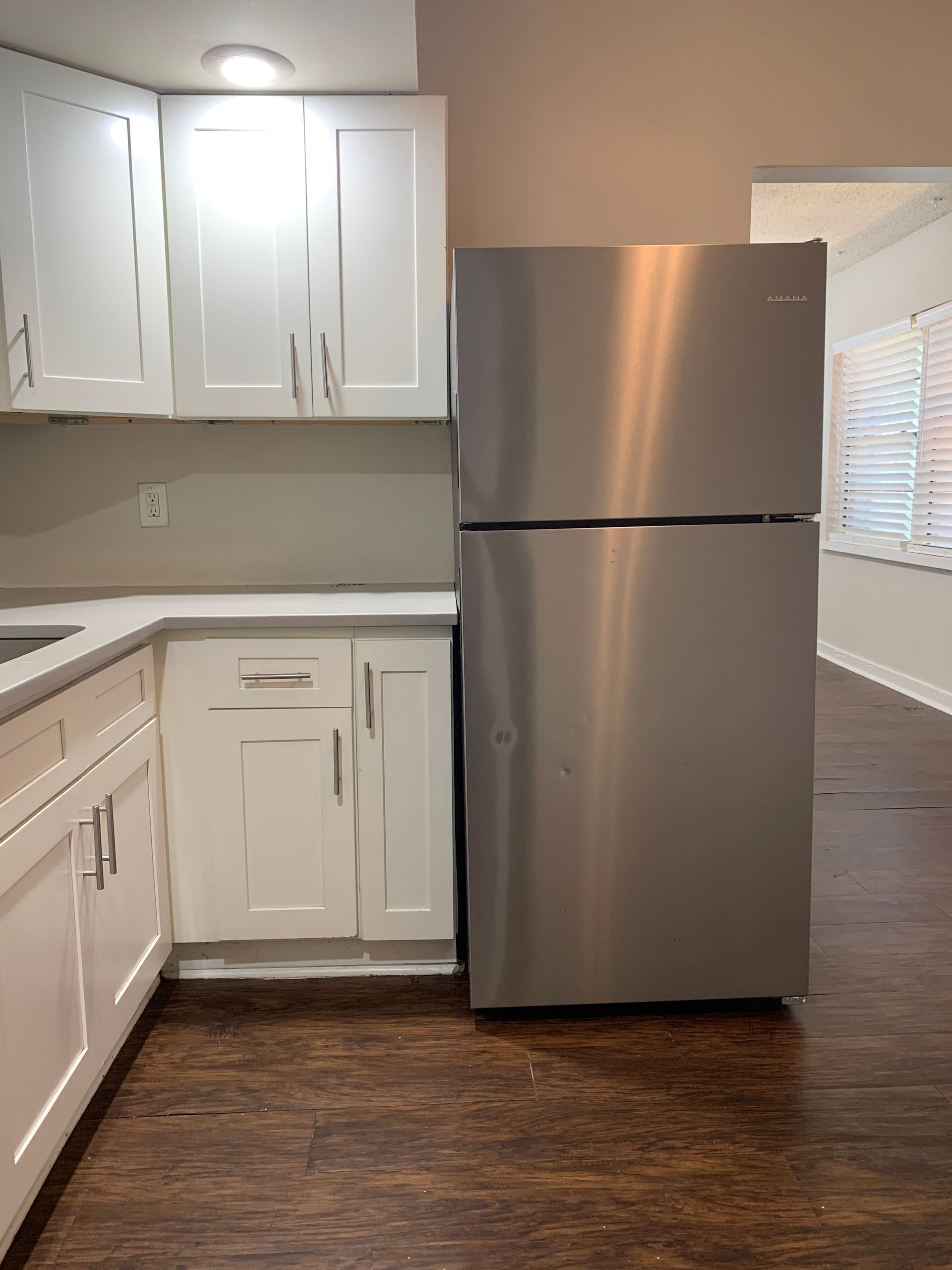 Stainless steel refrigerator next to white kitchen cabinets and countertop. Dark wood floor.