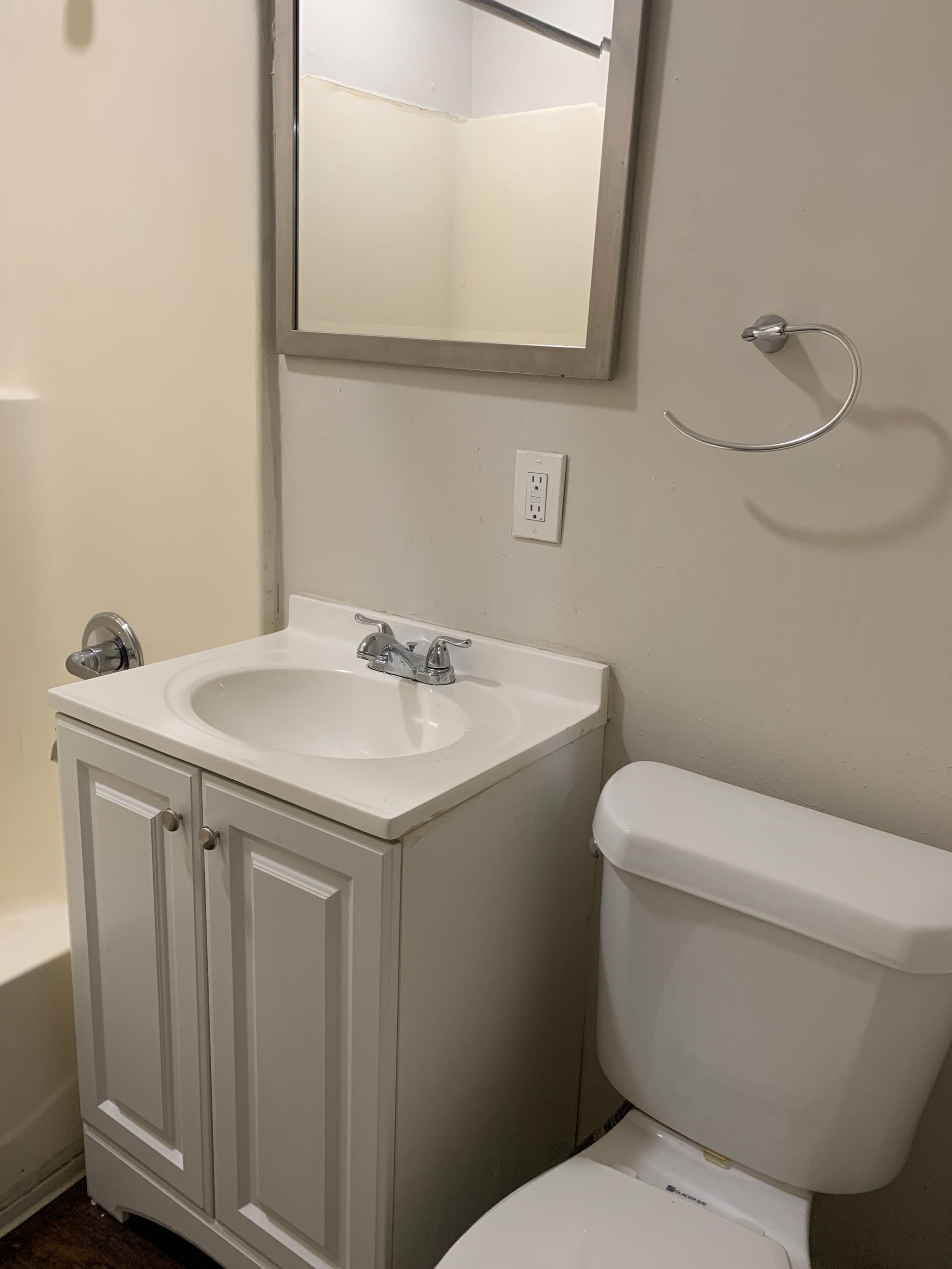 A bathroom with a white vanity, toilet, mirror, and towel ring against a beige wall.