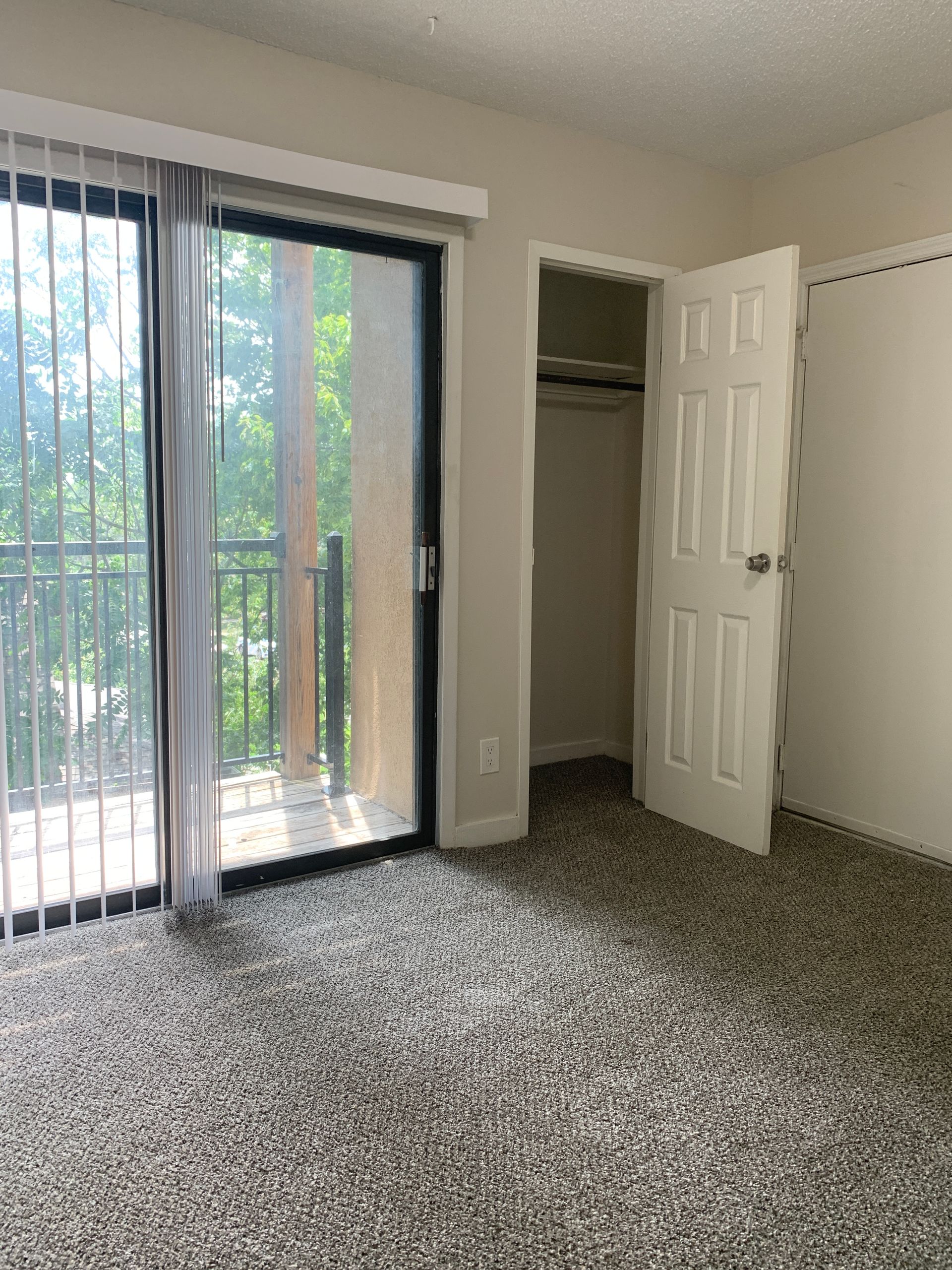 An empty bedroom with a sliding glass door to a balcony and a closet. Gray patterned carpet.