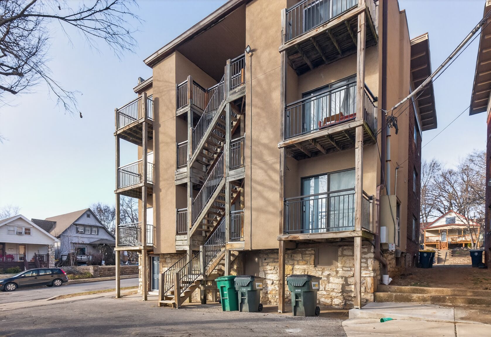Multi-story apartment building with exterior metal stairwell and balconies, beige exterior, and trash bins.