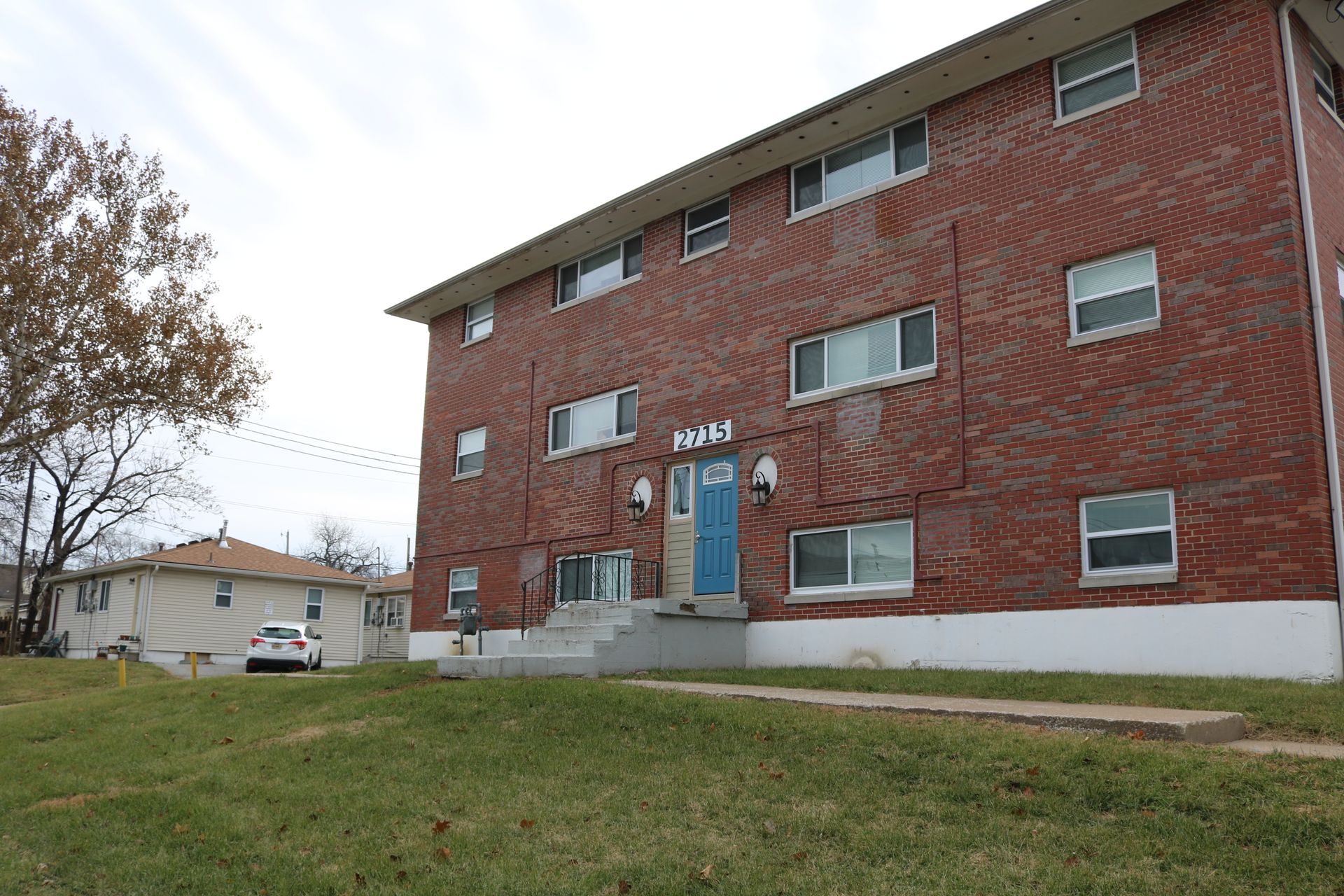 Brick apartment building with blue door, white trim, and a small yard on a cloudy day.