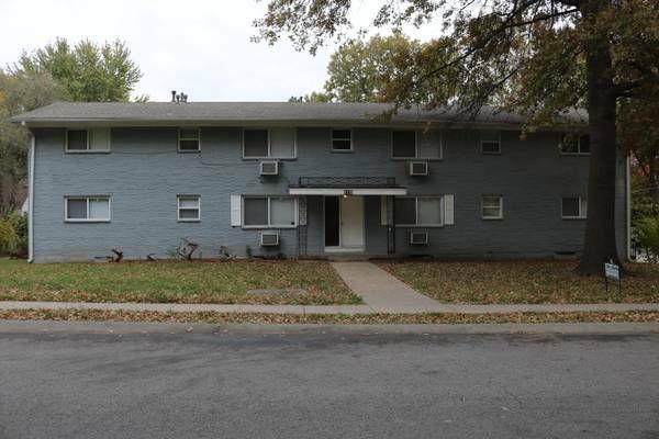Two-story apartment building with a blue-gray exterior, a central entrance, and a sidewalk leading to the street.