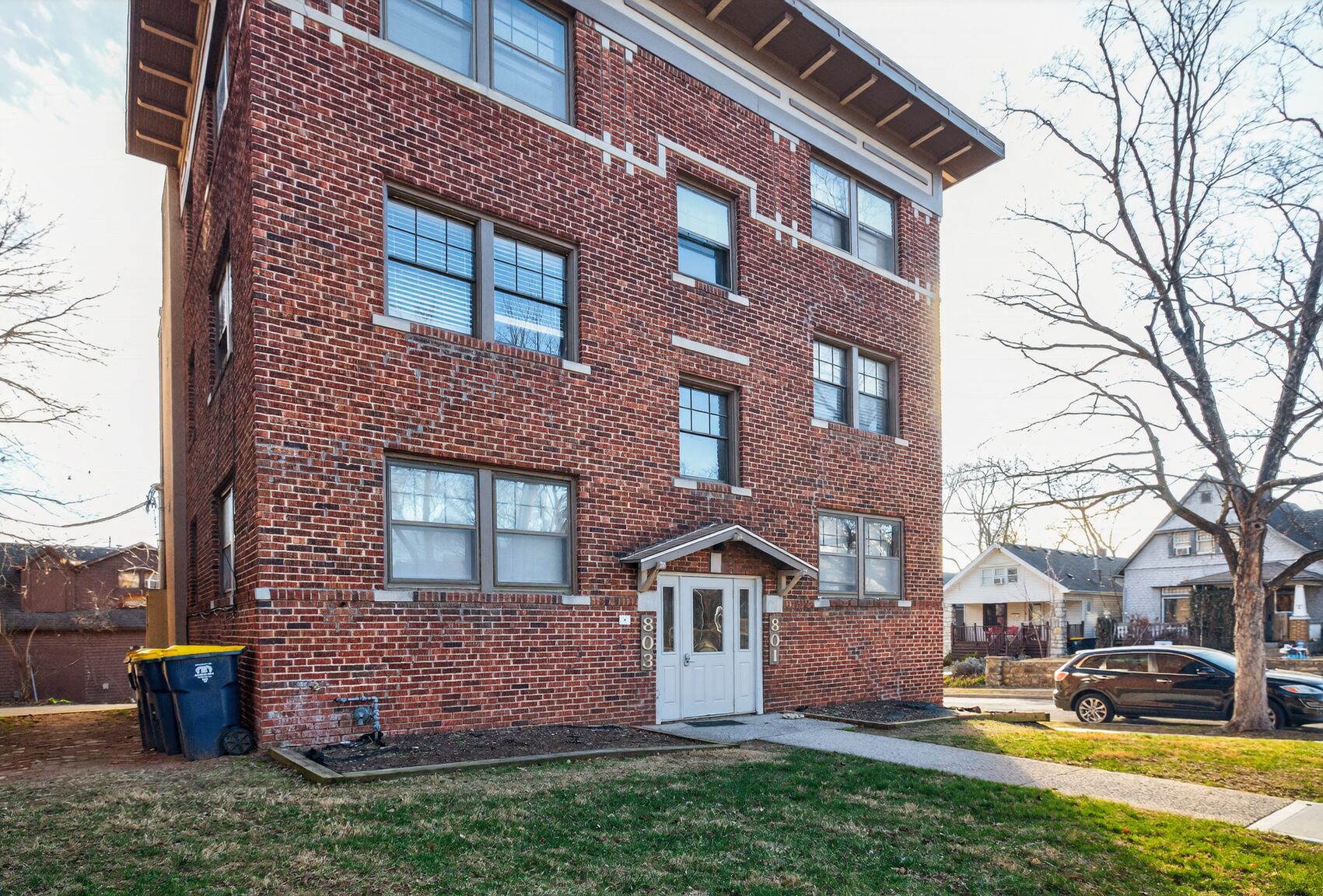 Brick apartment building with entrance, grass, trees, and parked cars.