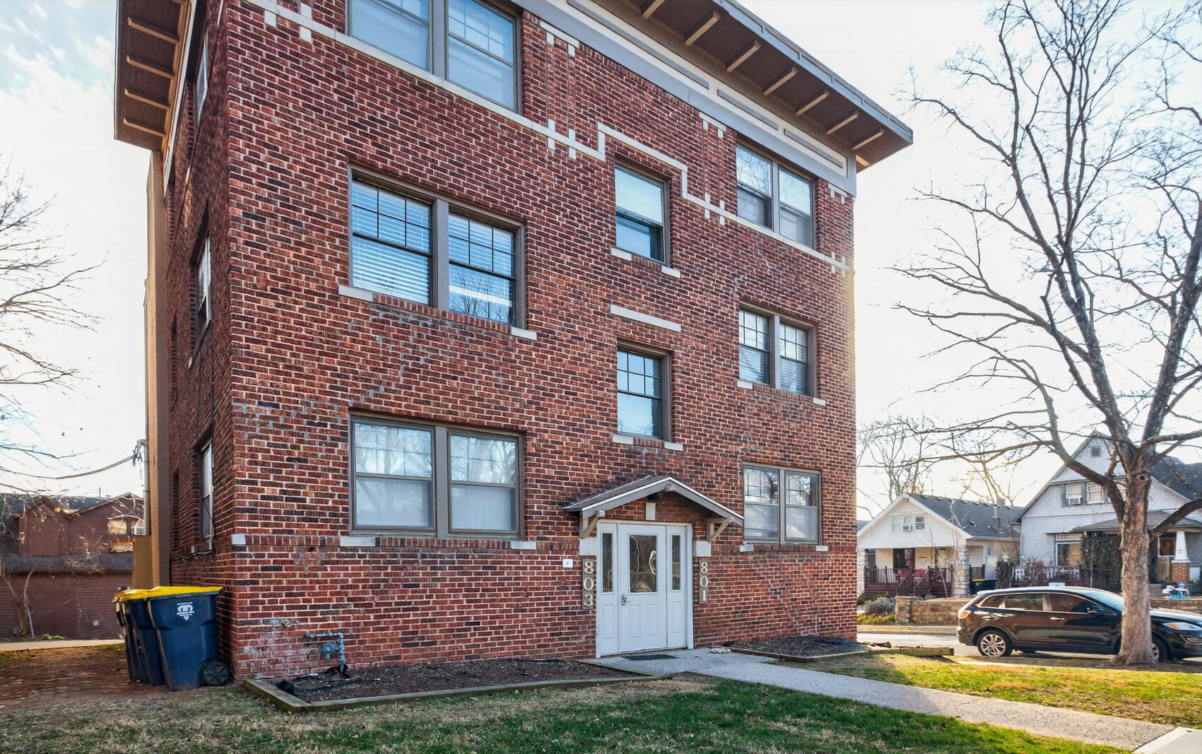 Brick apartment building with entrance, grass, trees, and parked cars.