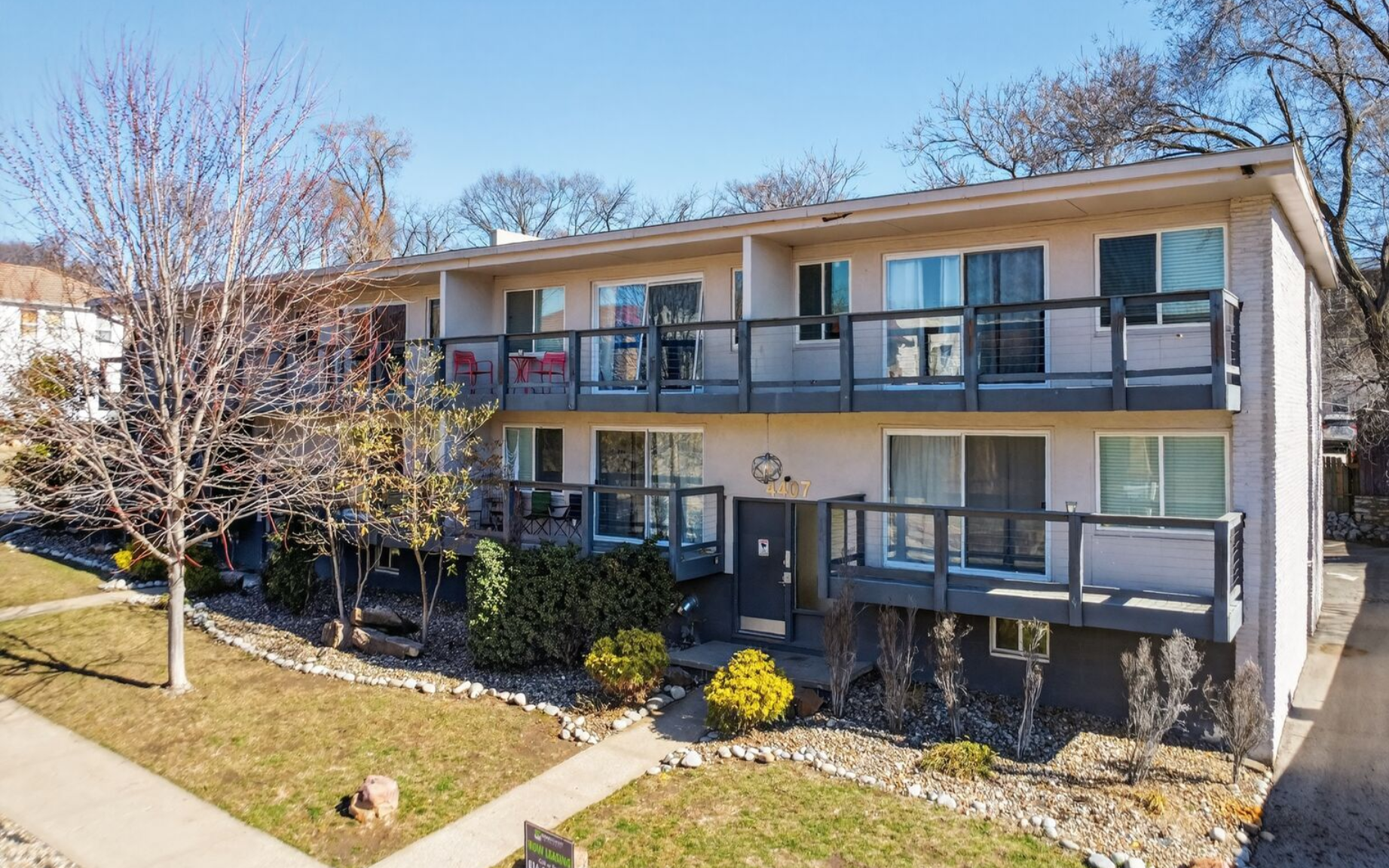 A two-story apartment building with beige siding and dark balconies, surrounded by dormant trees and landscaping.