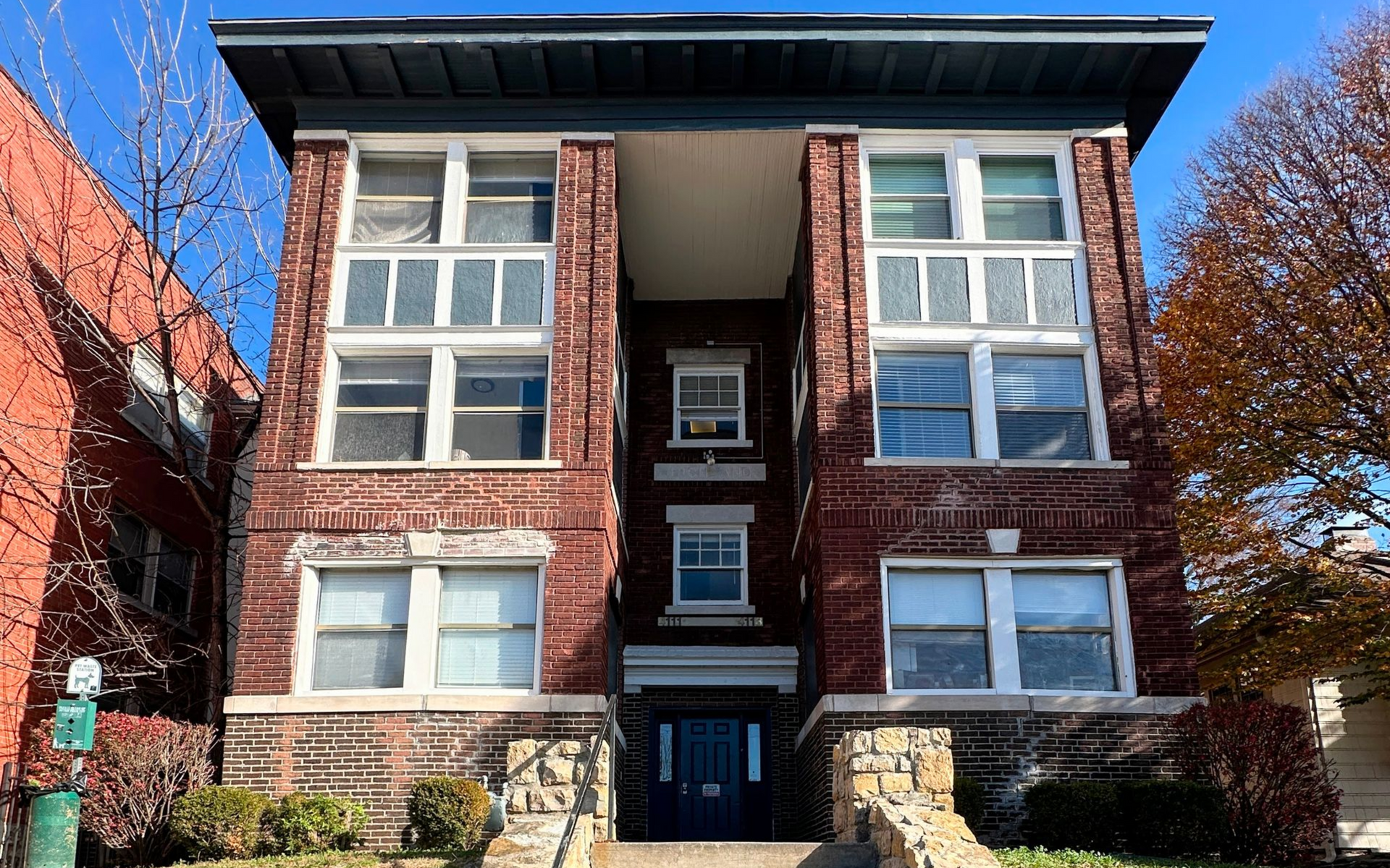 A three-story, red brick apartment building with a recessed center entrance, stone foundation, and a dark overhanging roof.
