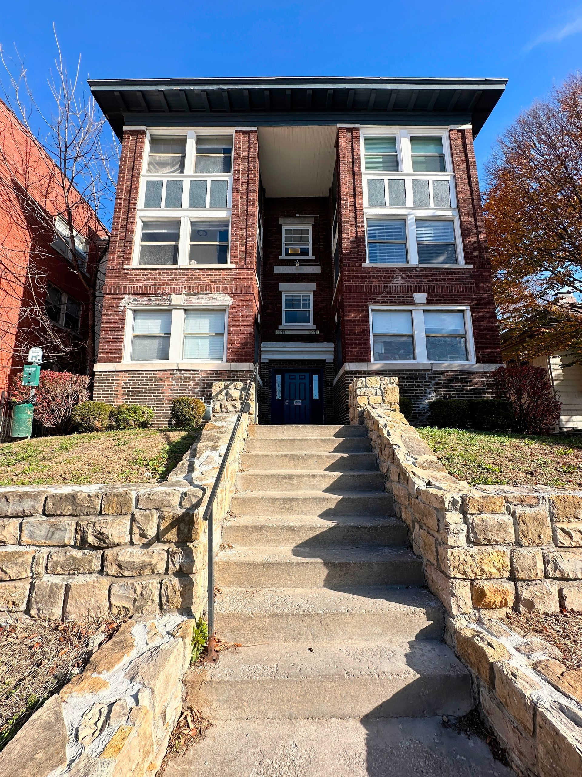 A red brick apartment building with a central entrance, reached by a wide stone staircase leading up from the sidewalk.