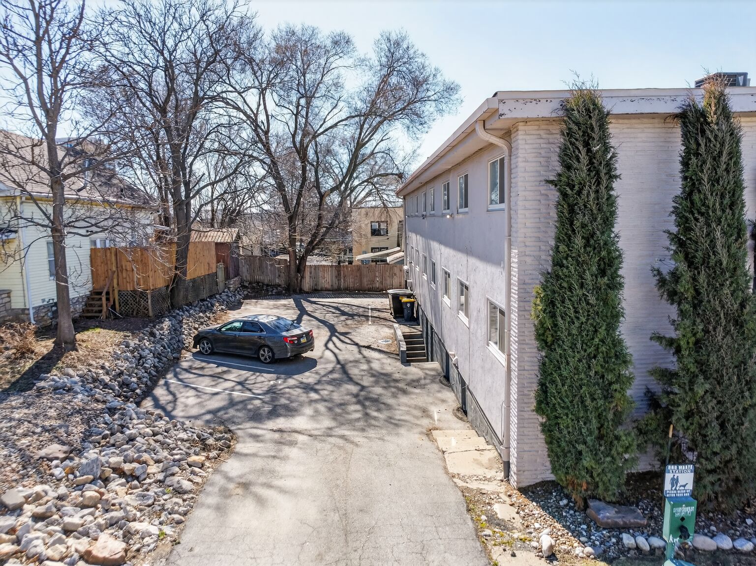 A gray car is parked in the driveway beside a multi-story, light-colored brick apartment building.