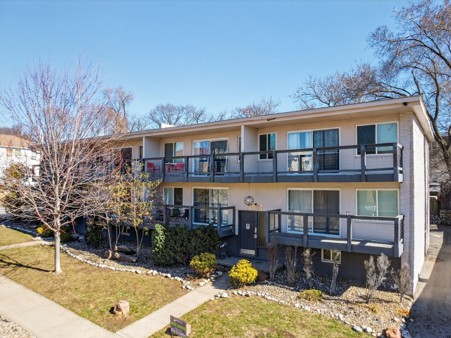 A two-story apartment complex with beige stucco walls, dark gray balconies, and bare trees on a sunny day.