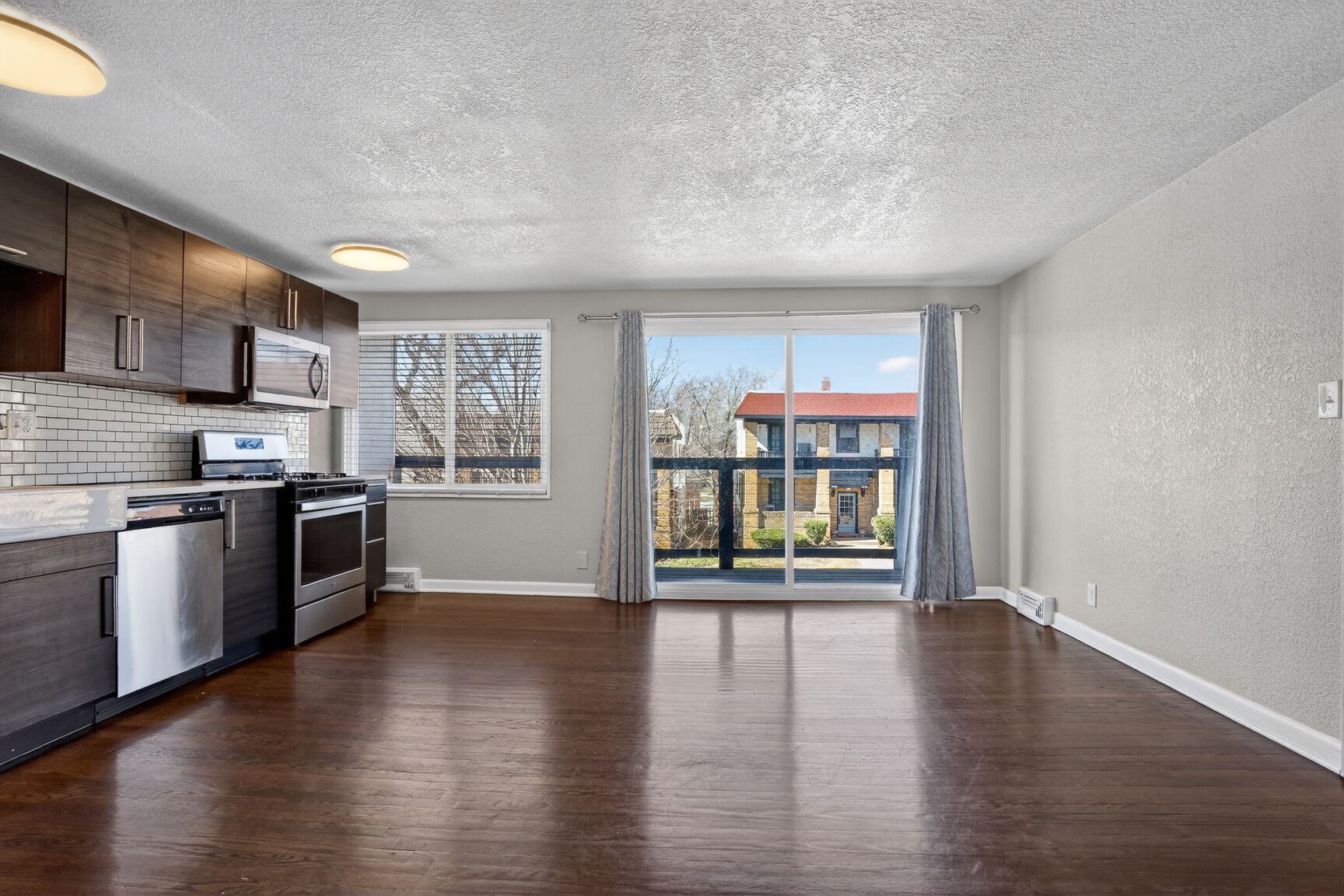 An open-concept kitchen and living area with dark brown floors, dark wood cabinets, stainless appliances, and a balcony.