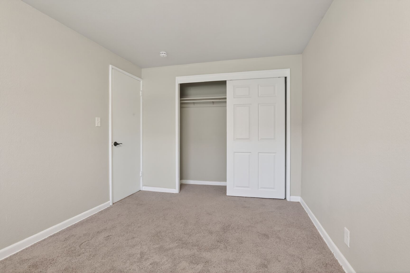 An empty bedroom with light gray walls, tan carpet, a white closed door, and an open white sliding closet door.