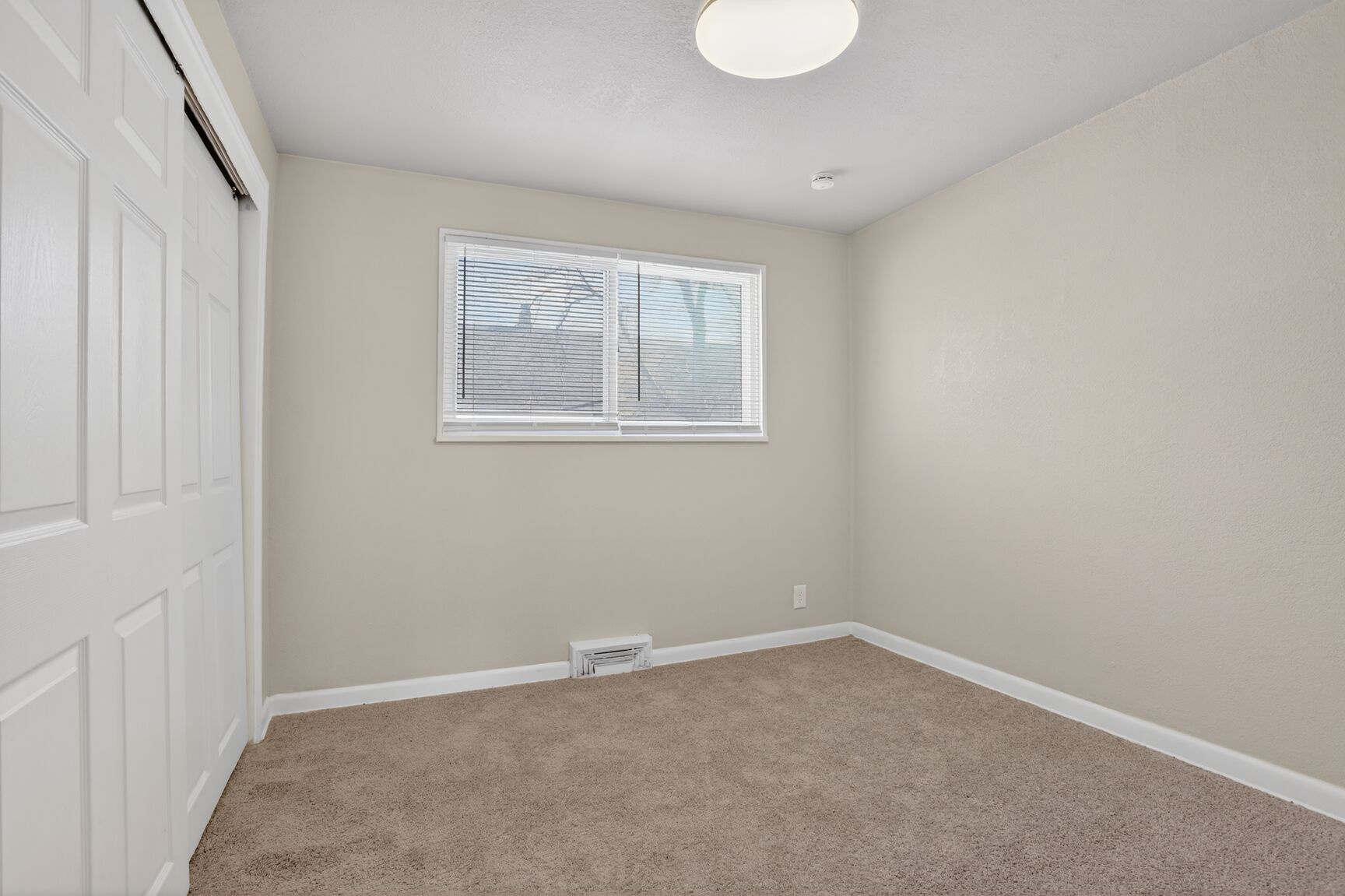 A vacant room with light-colored walls, carpeted flooring, a white closet, and a single window with closed blinds.