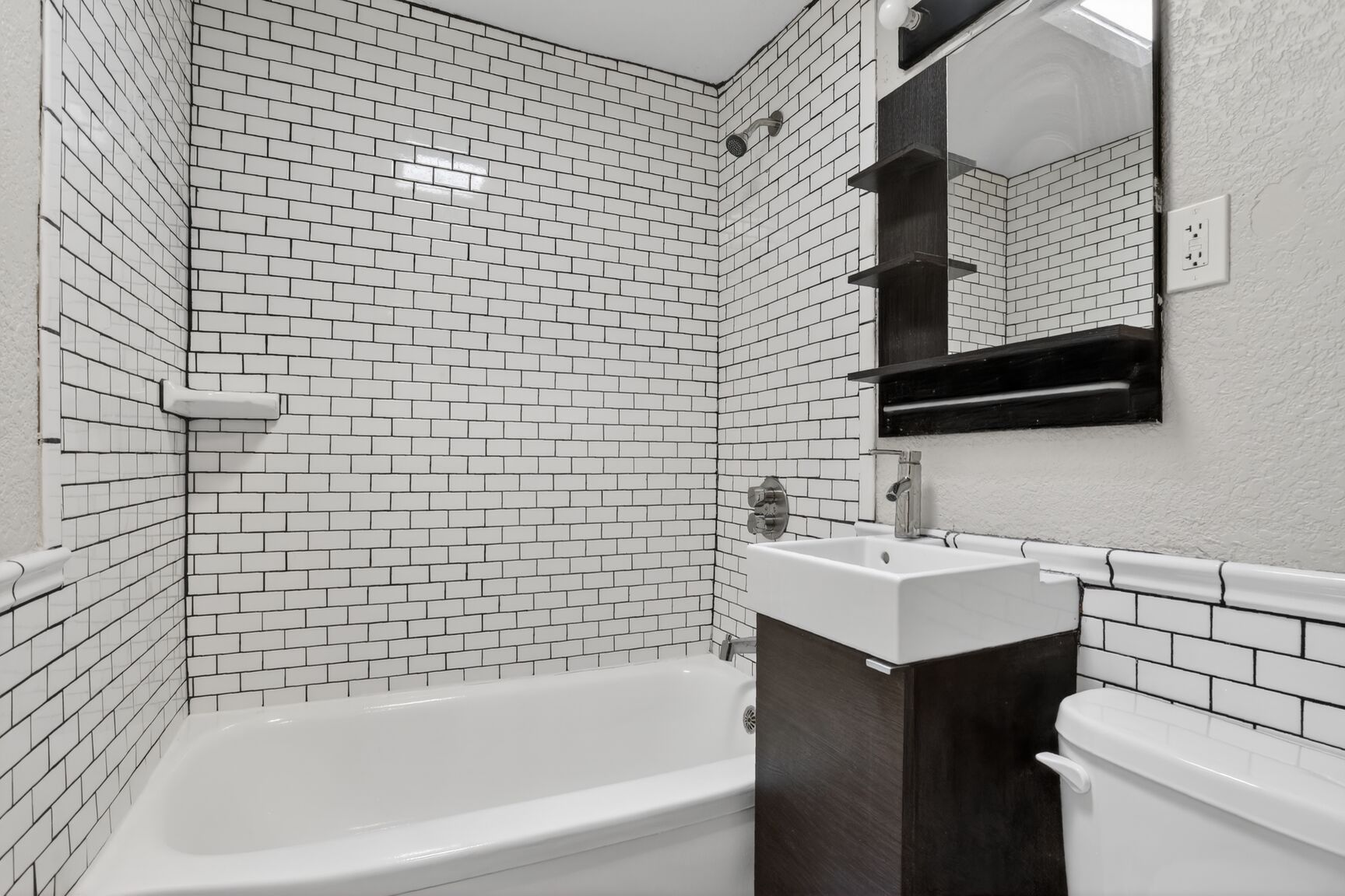 A small bathroom with white subway tile walls, a white bathtub, a dark wood vanity with a white sink, and a black mirror.