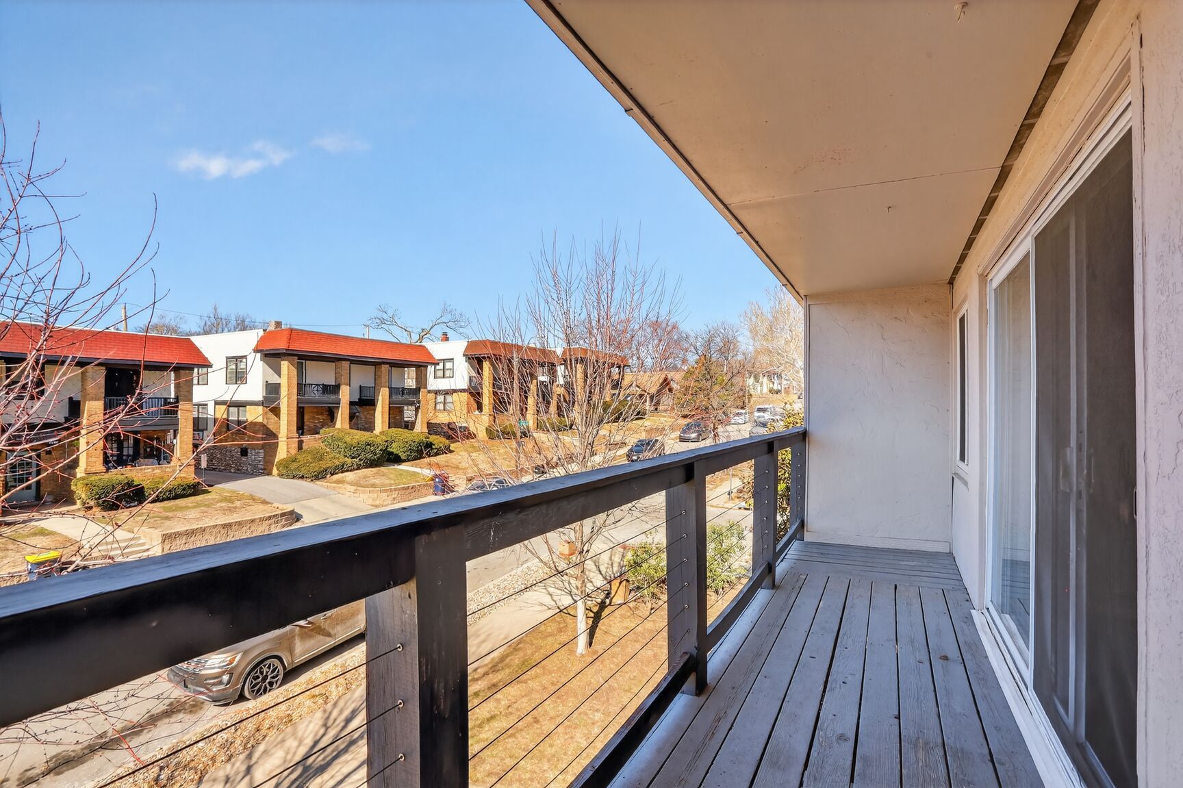 A view from a wooden apartment balcony overlooking a street, sparse trees, and neighboring townhomes under a clear sky.