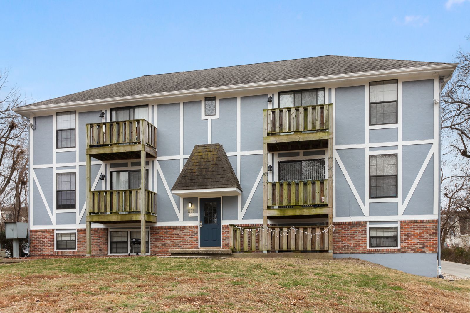 Two-story light blue apartment building with wooden balconies, brown roof, and brick facade.