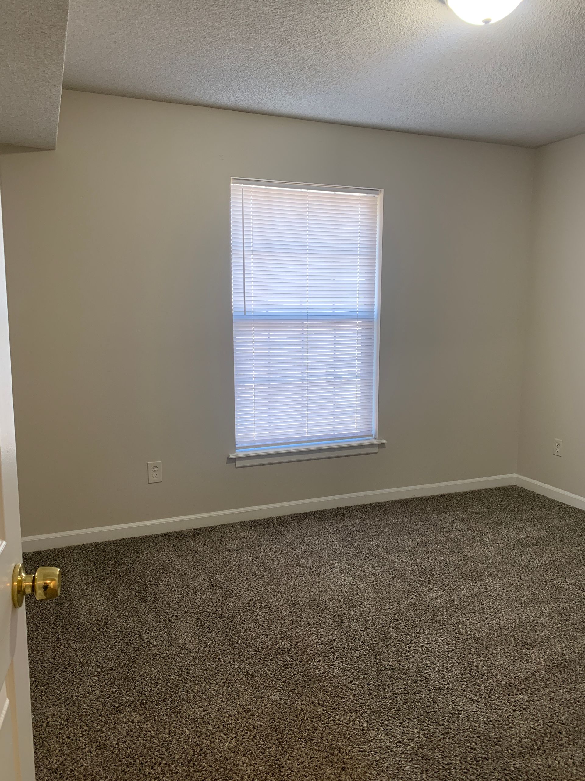 Empty room with tan walls, a window with blinds, and a brown speckled carpet.