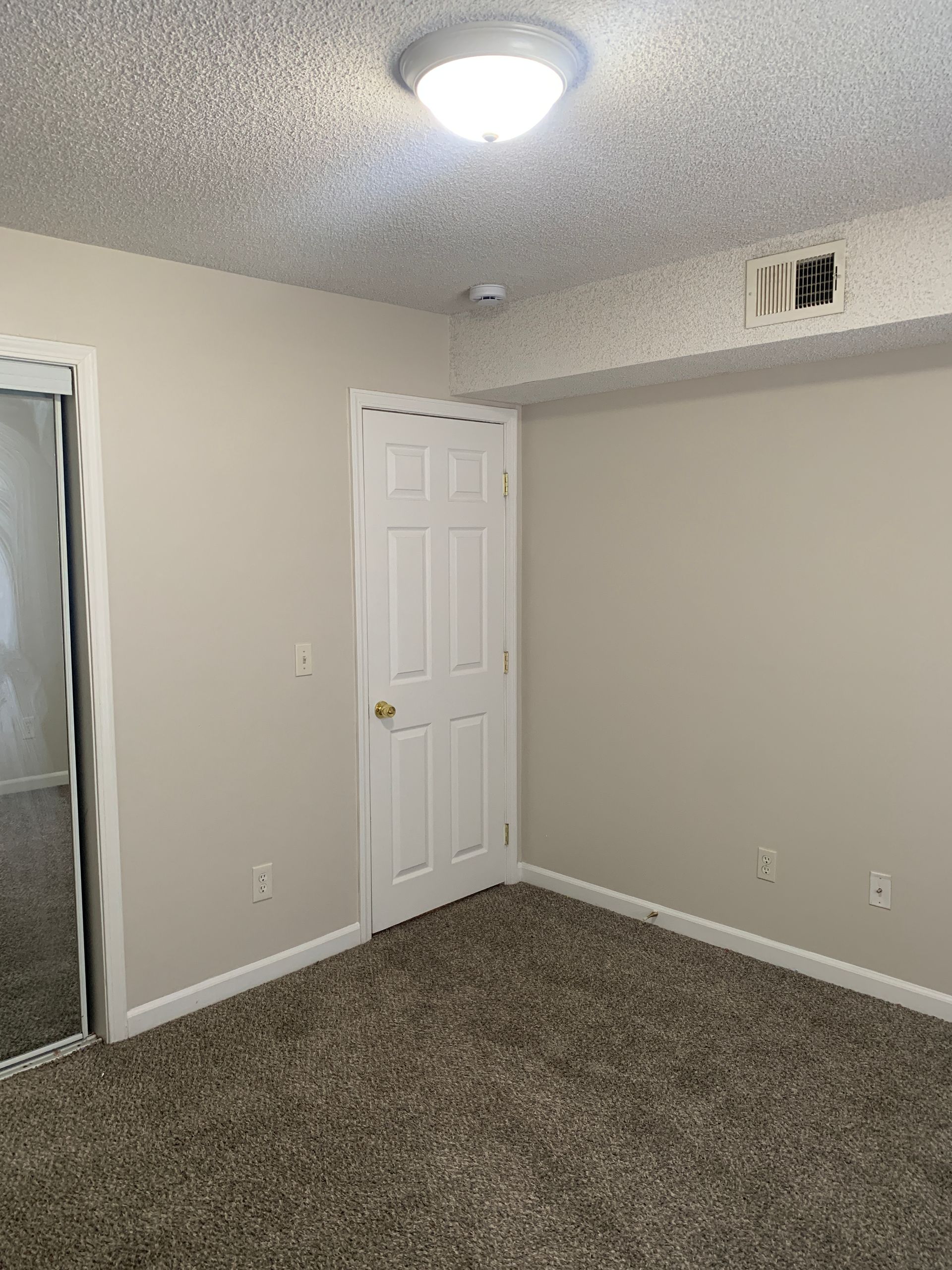 Empty bedroom with tan walls, brown carpet, white door and ceiling light.
