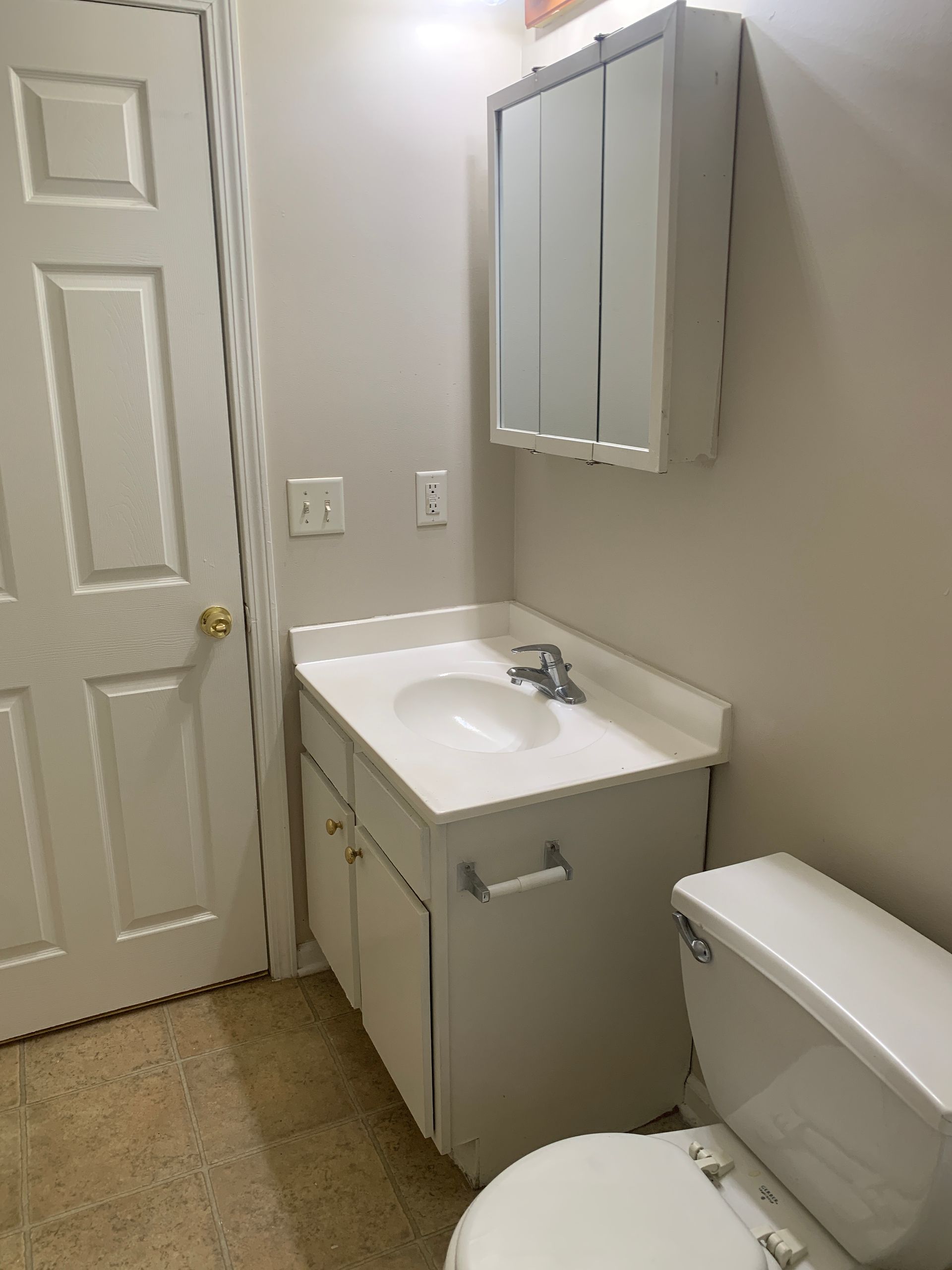 Small bathroom with white vanity, toilet, and medicine cabinet; tan flooring.