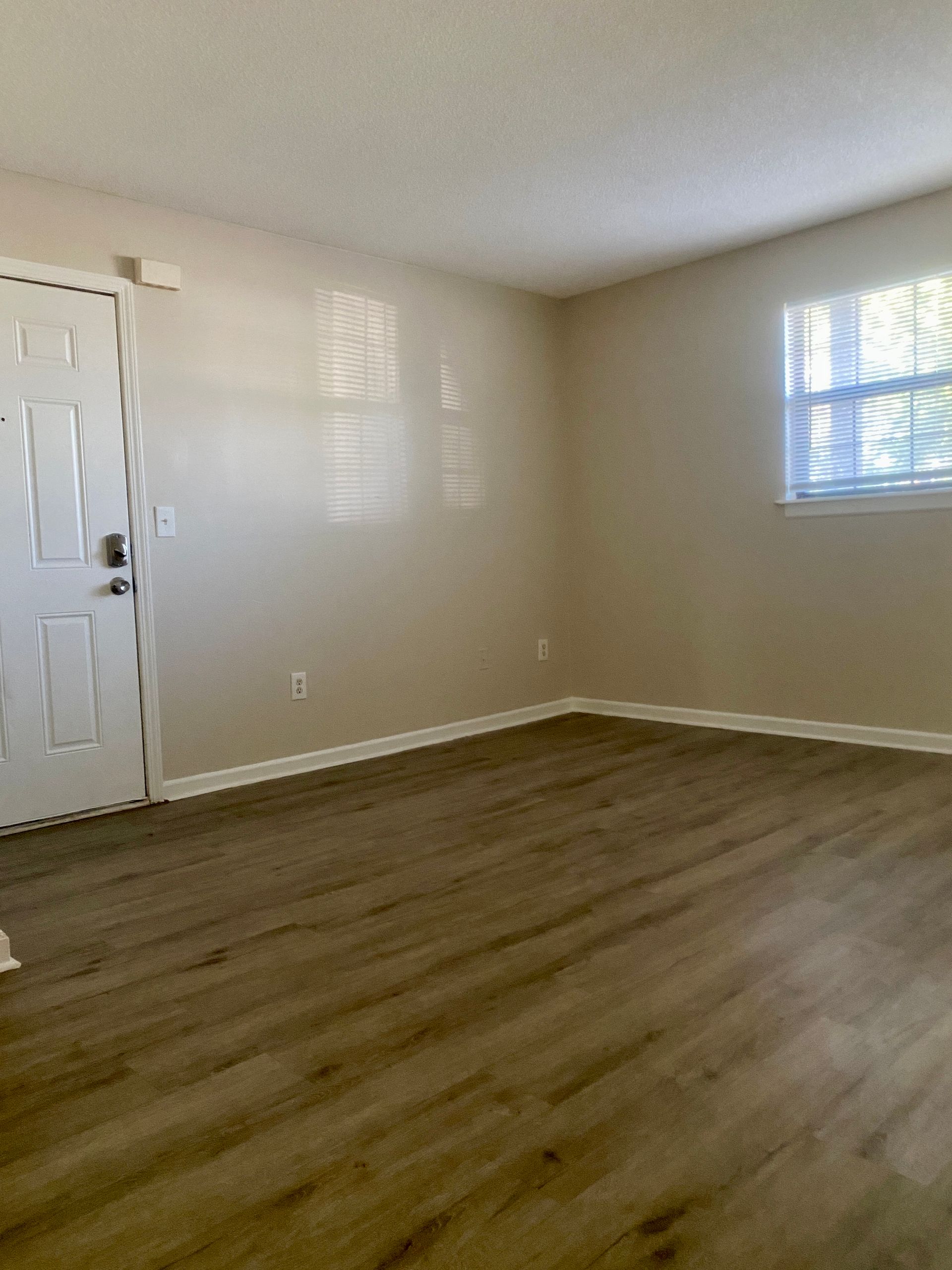 Empty room with wood-look flooring, white door and trim, two windows with blinds, and beige walls.
