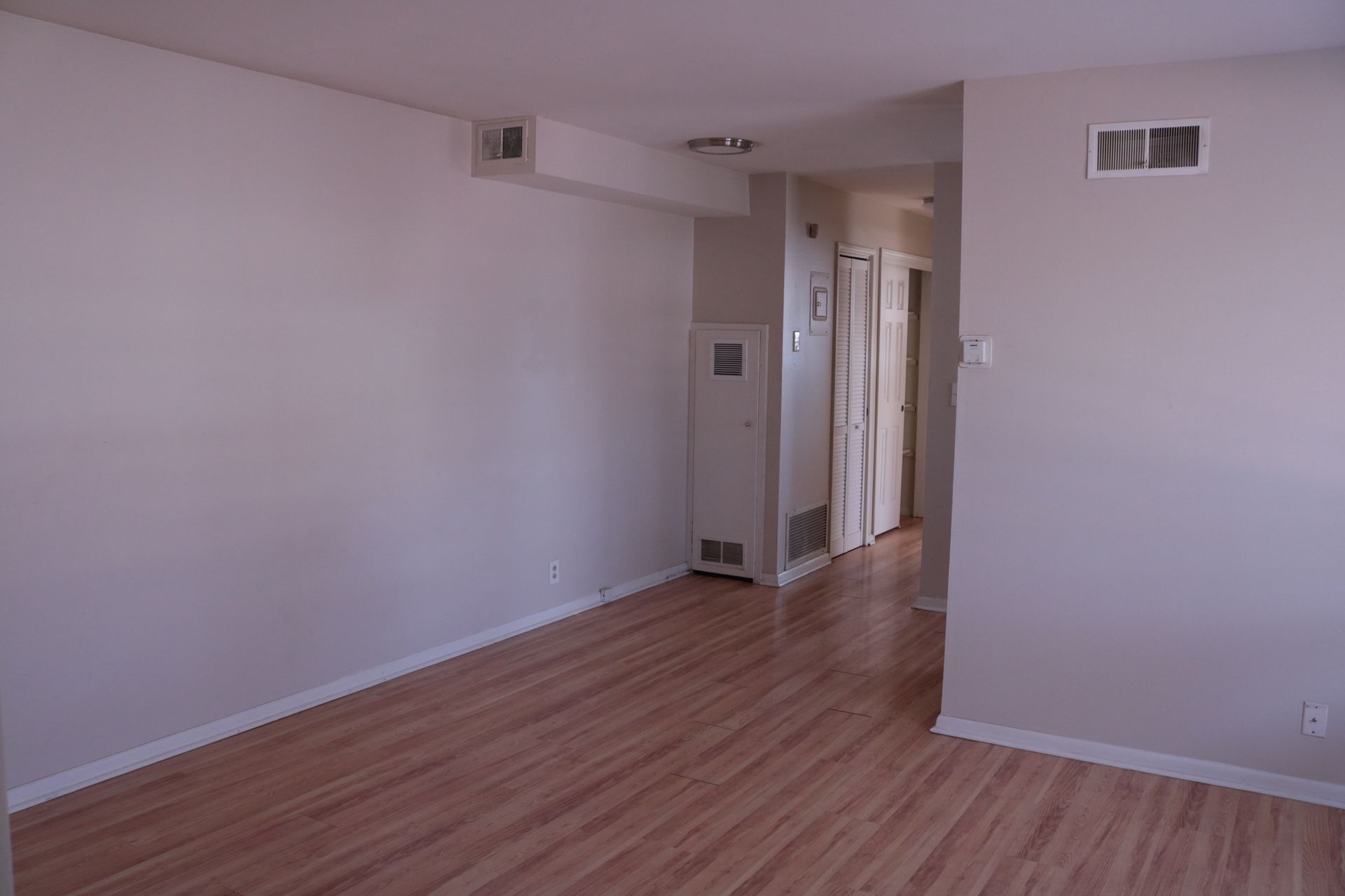Empty living room with light wooden floors, white walls, and an entryway with doors.